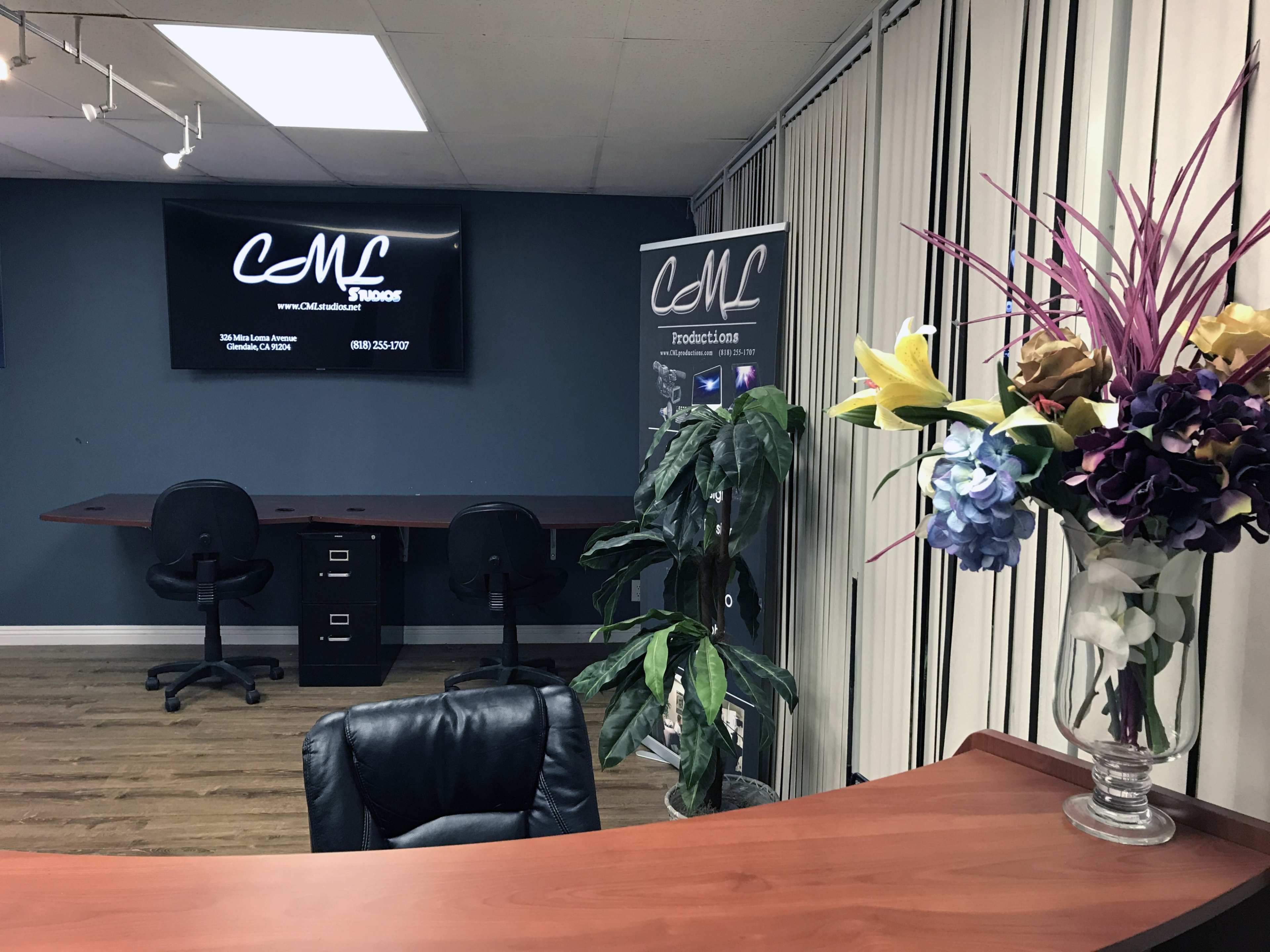 An office space featuring a reception desk, a potted plant, a television screen on the wall, and a promotional banner alongside a decorative flower arrangement.