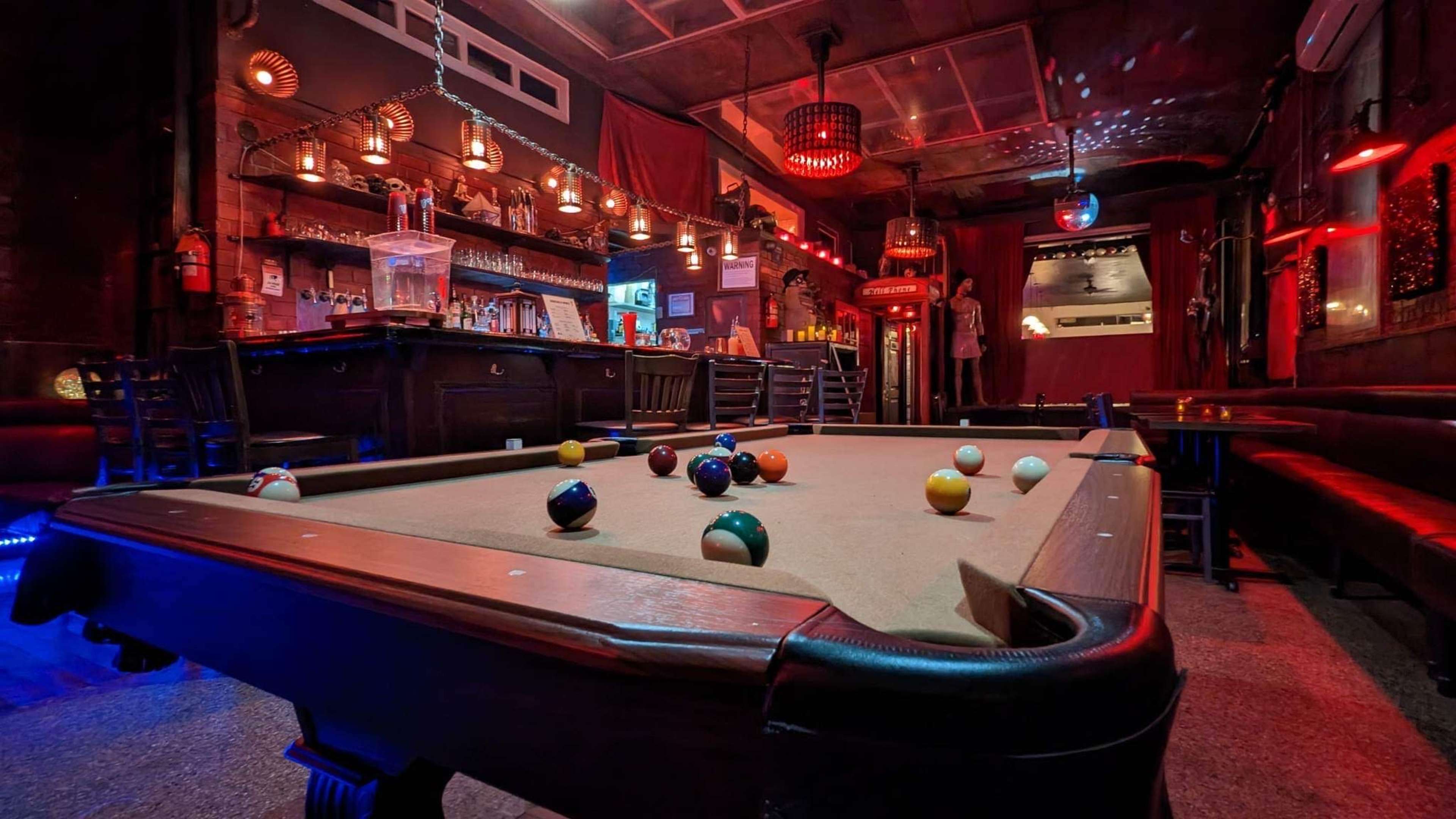 A dimly lit bar features a pool table with colorful balls scattered on its surface, surrounded by shelves of alcohol and red lighting.