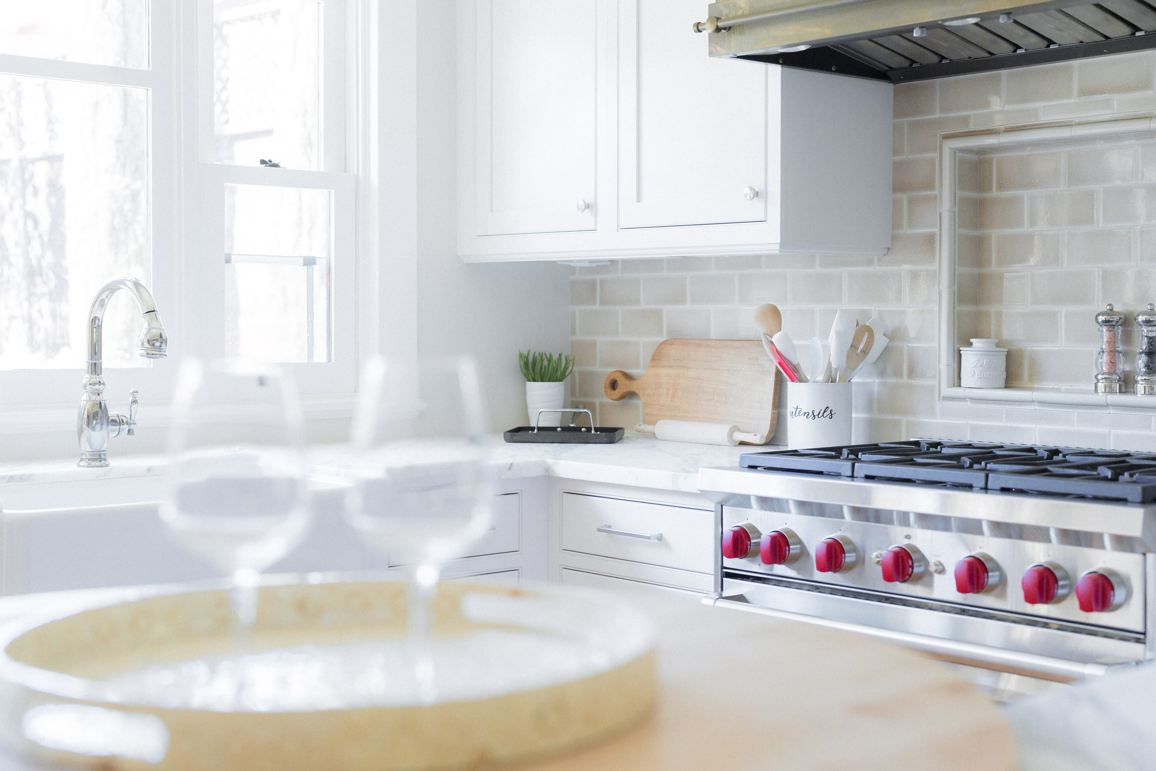 A modern kitchen features white cabinetry, a stainless steel stove with red knobs, and a marble countertop with glassware and a wooden cutting board.