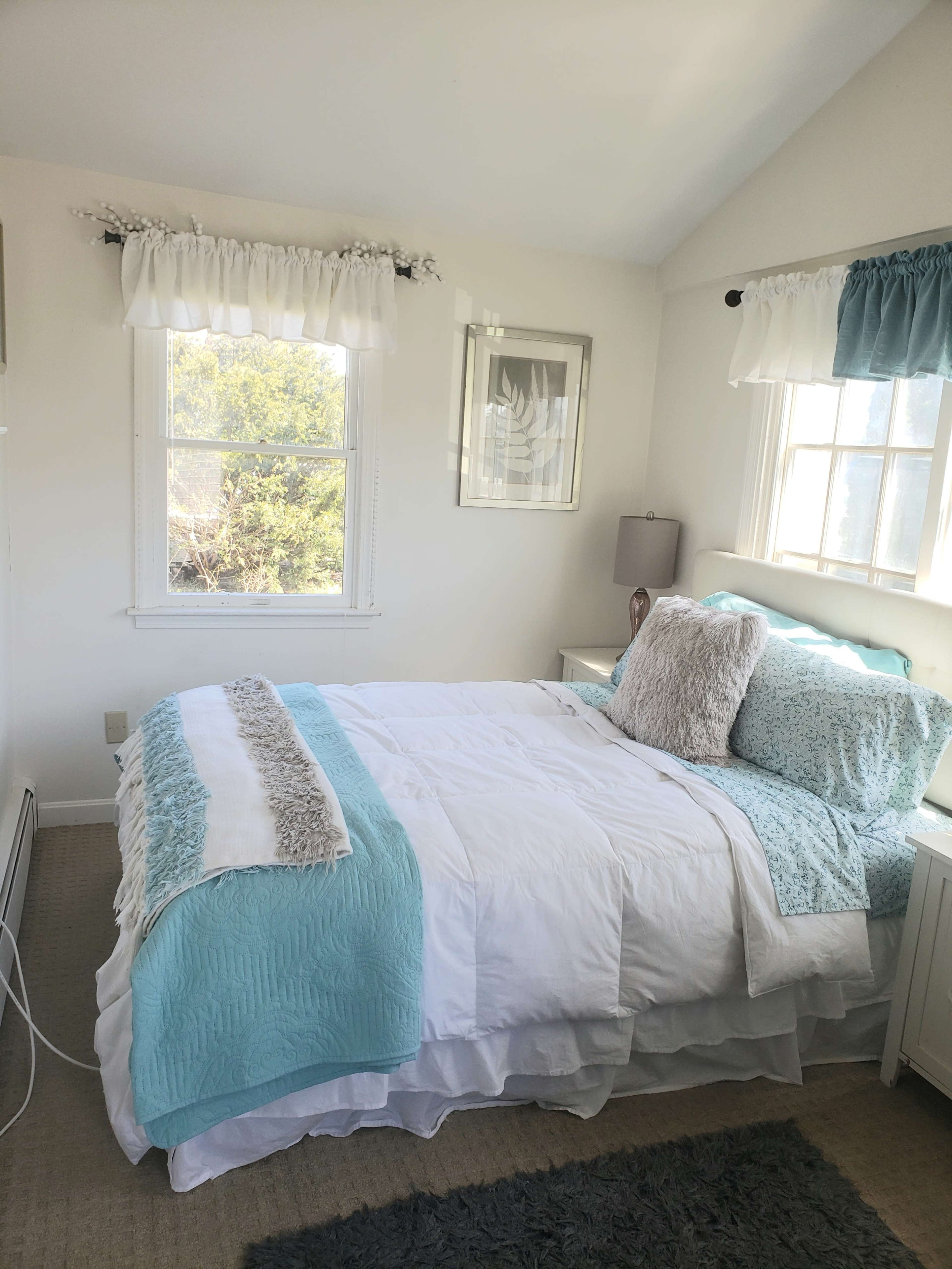 A neatly arranged bedroom with a white bedspread, light blue accents, and two windows that let in natural light.