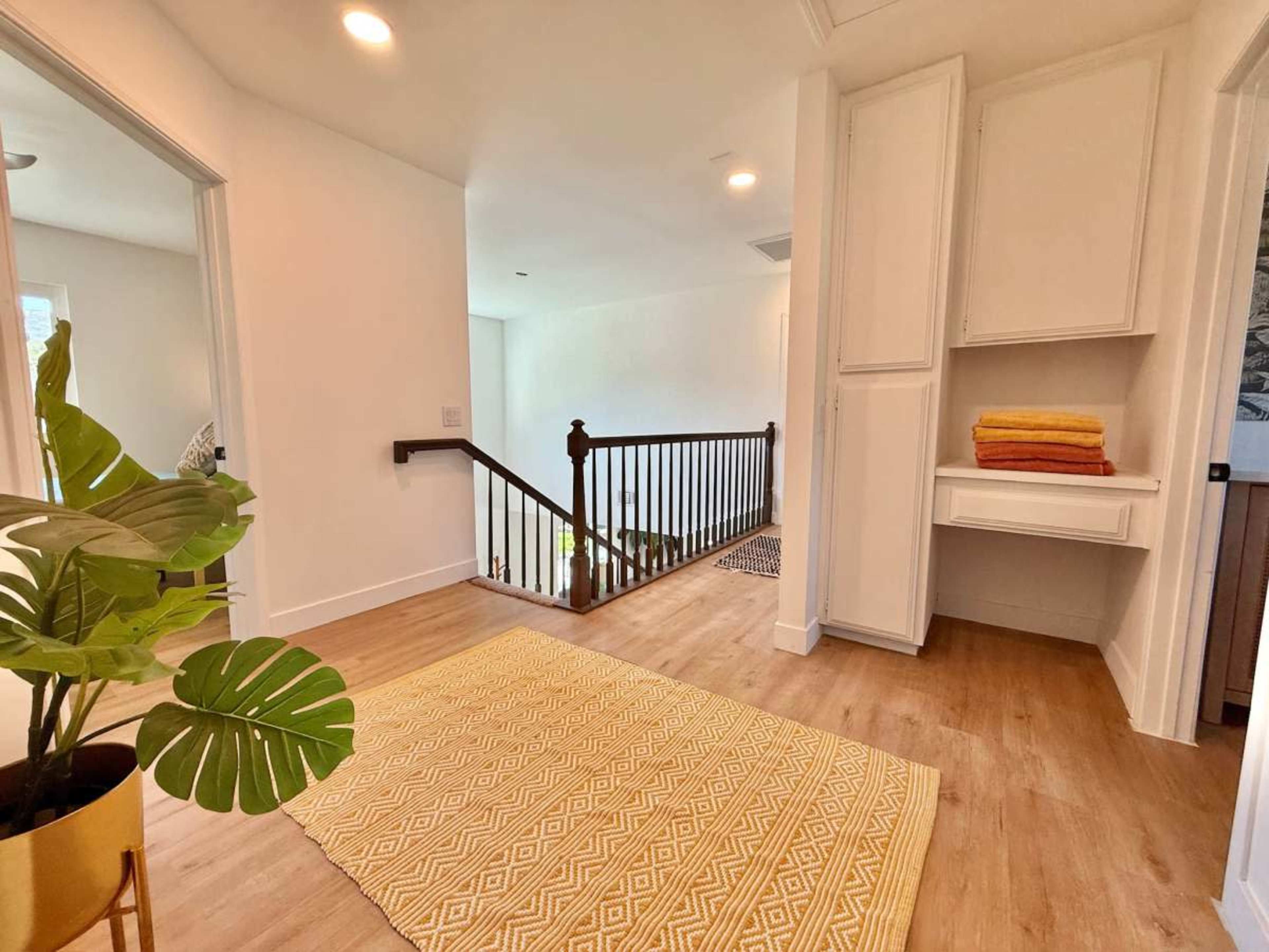The image shows a bright hallway with wooden flooring, a staircase with a dark railing, and a small nook with stacked towels and a potted plant.