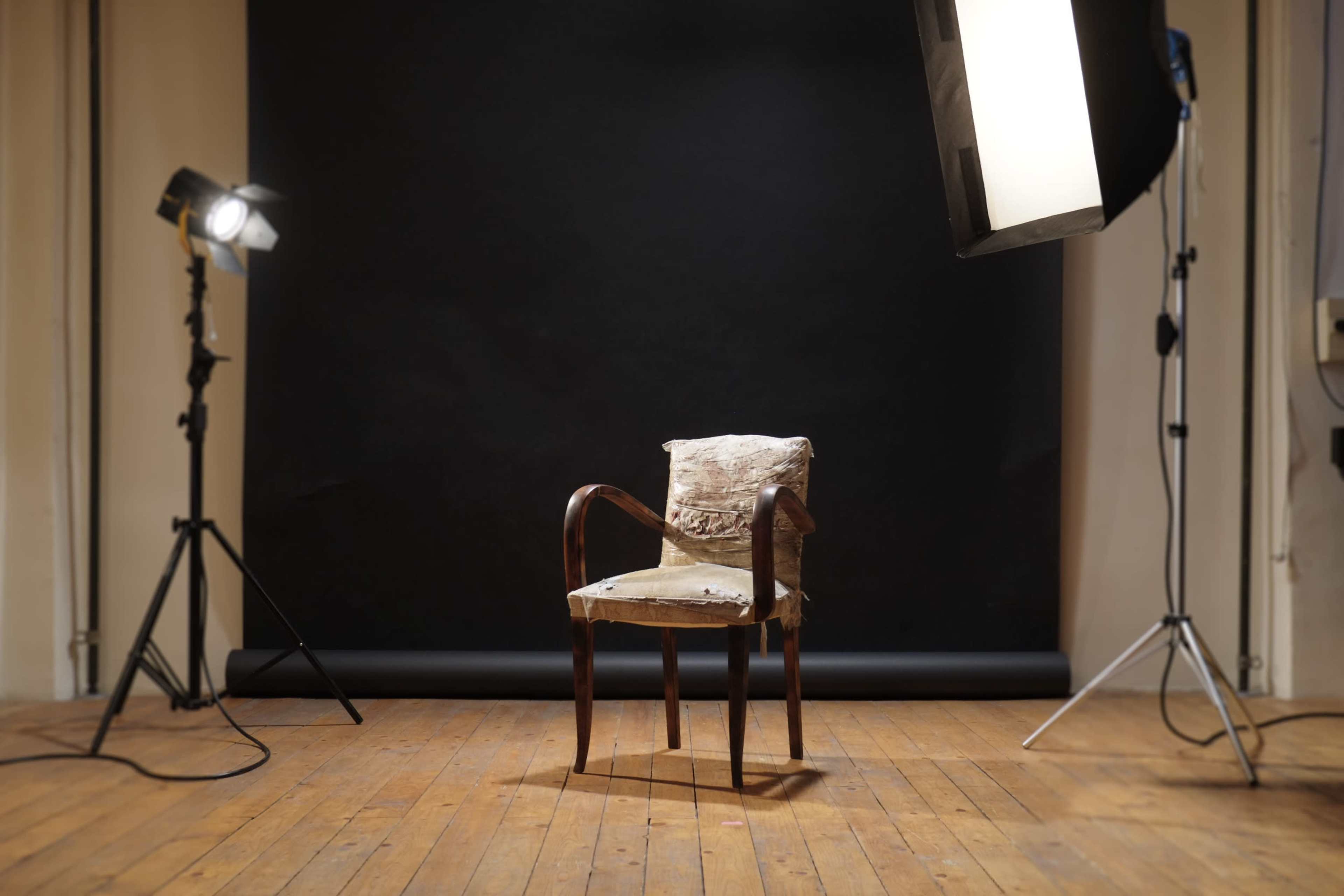 A worn chair is placed in front of a black backdrop, illuminated by studio lights on either side.