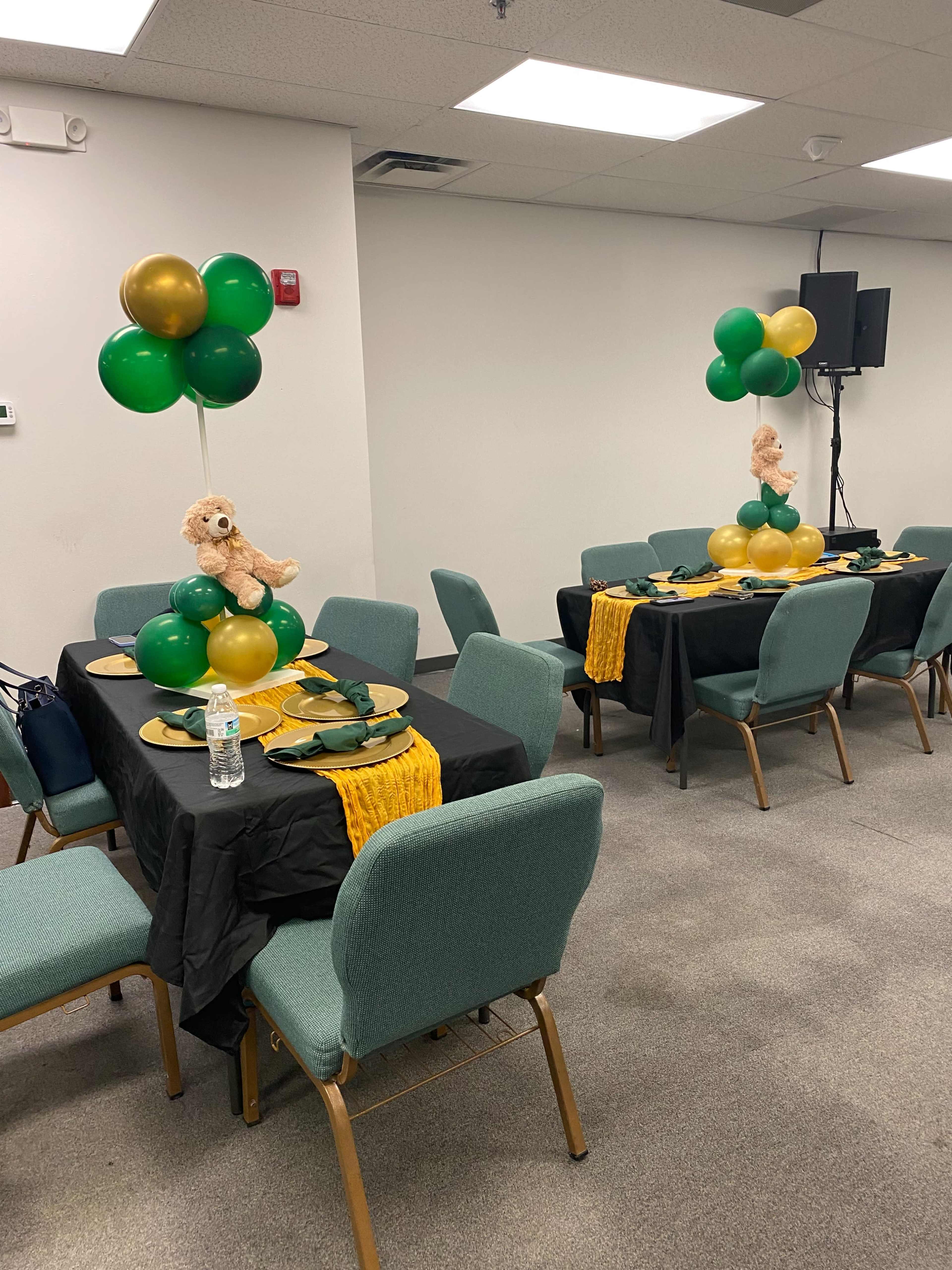 The image shows a decorated banquet space with two tables set with black tablecloths, green and yellow balloons, and teddy bears as centerpieces.
