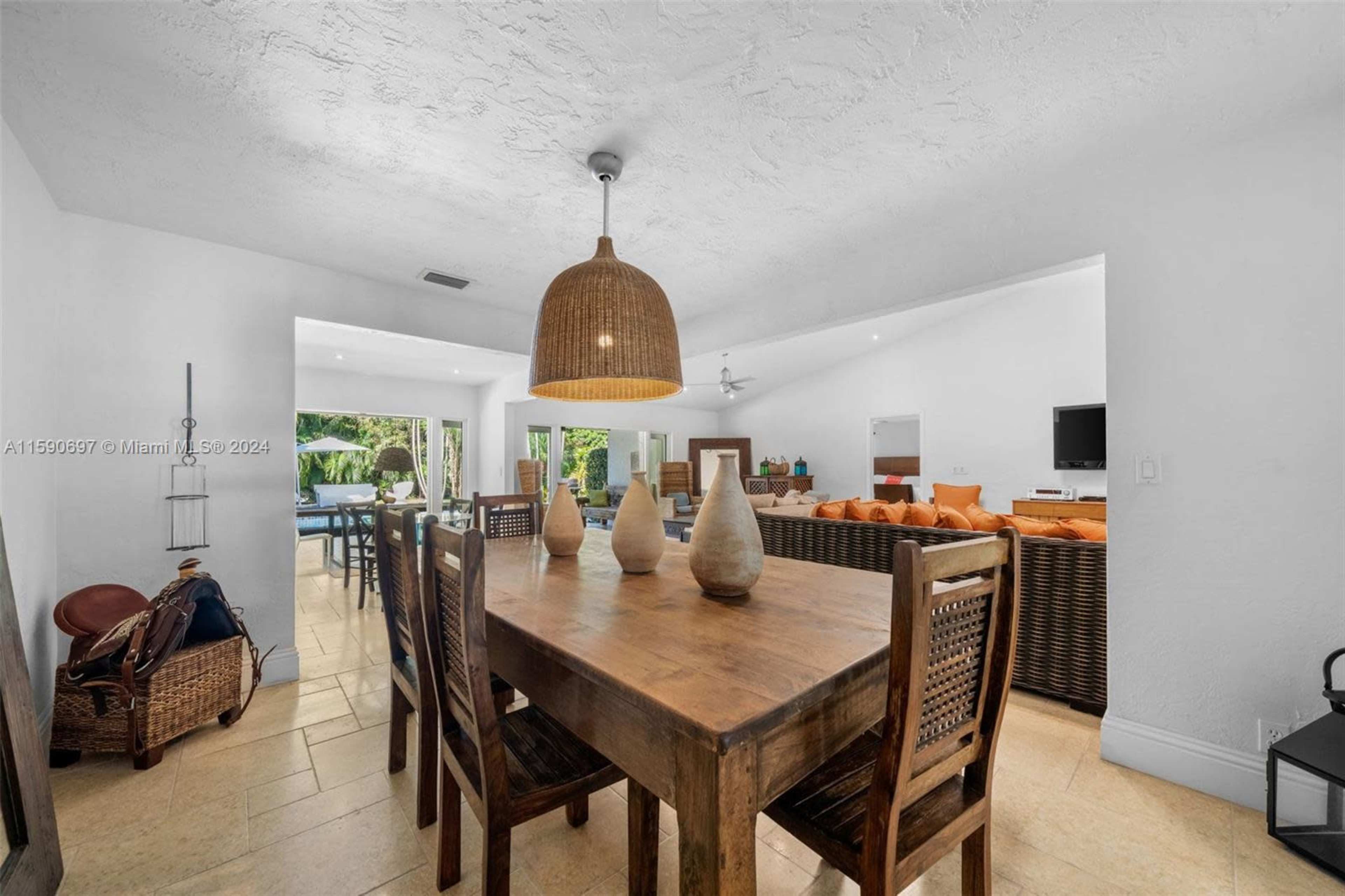 The image shows a dining area featuring a wooden table surrounded by chairs, with a view of a living space and outdoor pool area in the background.