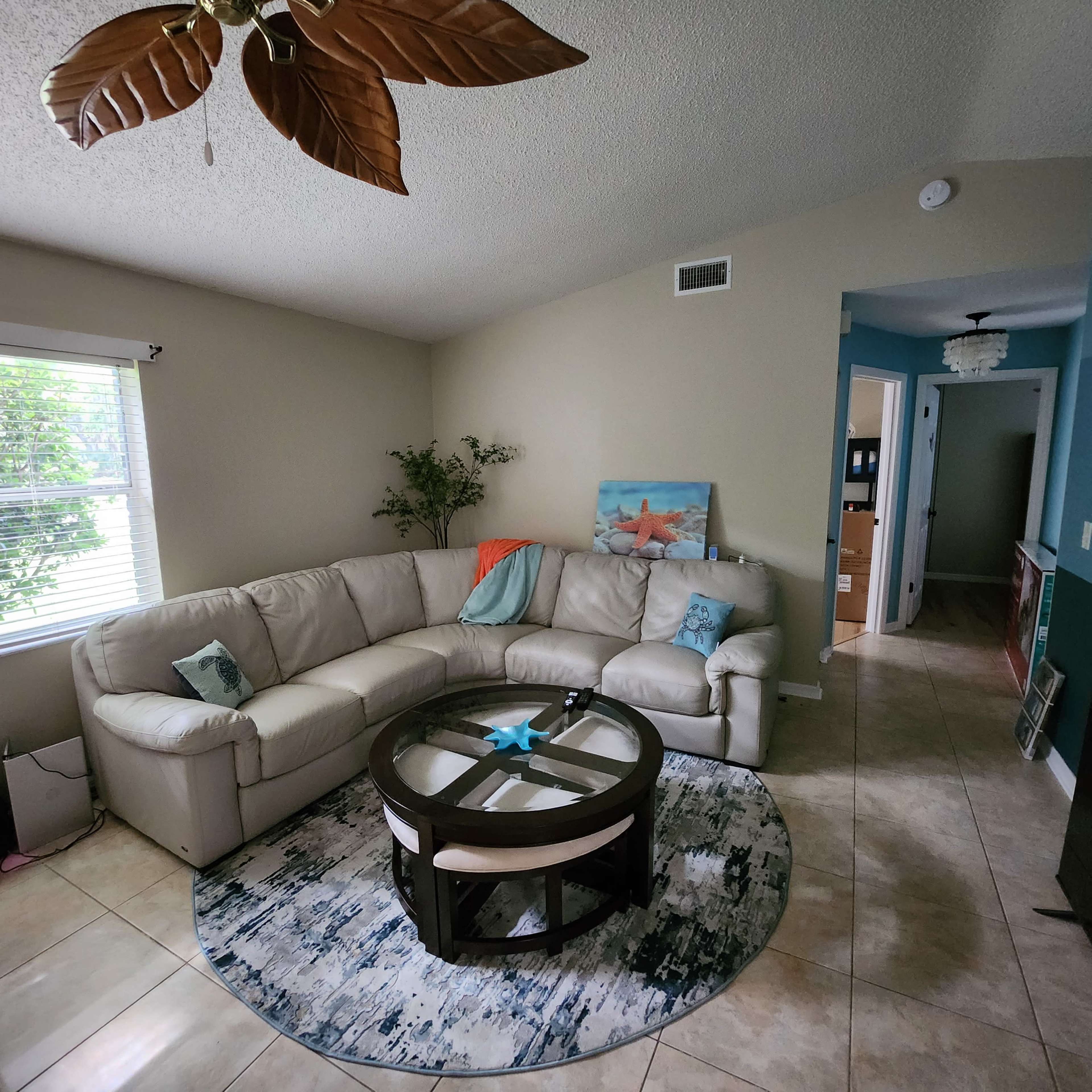 A light-colored sectional sofa arranged around a circular coffee table on a patterned rug in a living room with a ceiling fan and a small plant.