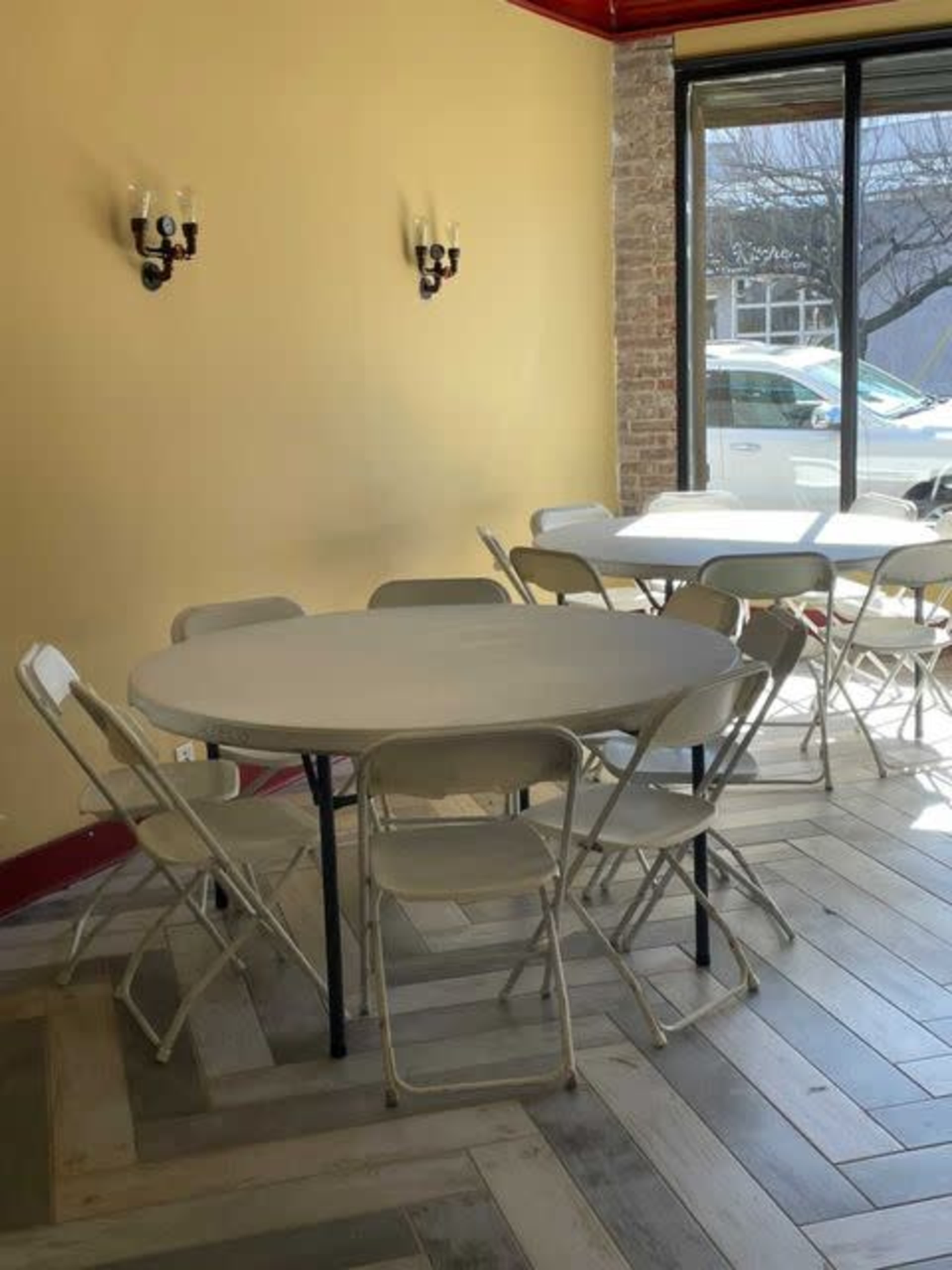 A round table surrounded by folding chairs is positioned in a brightly lit room with a large window.