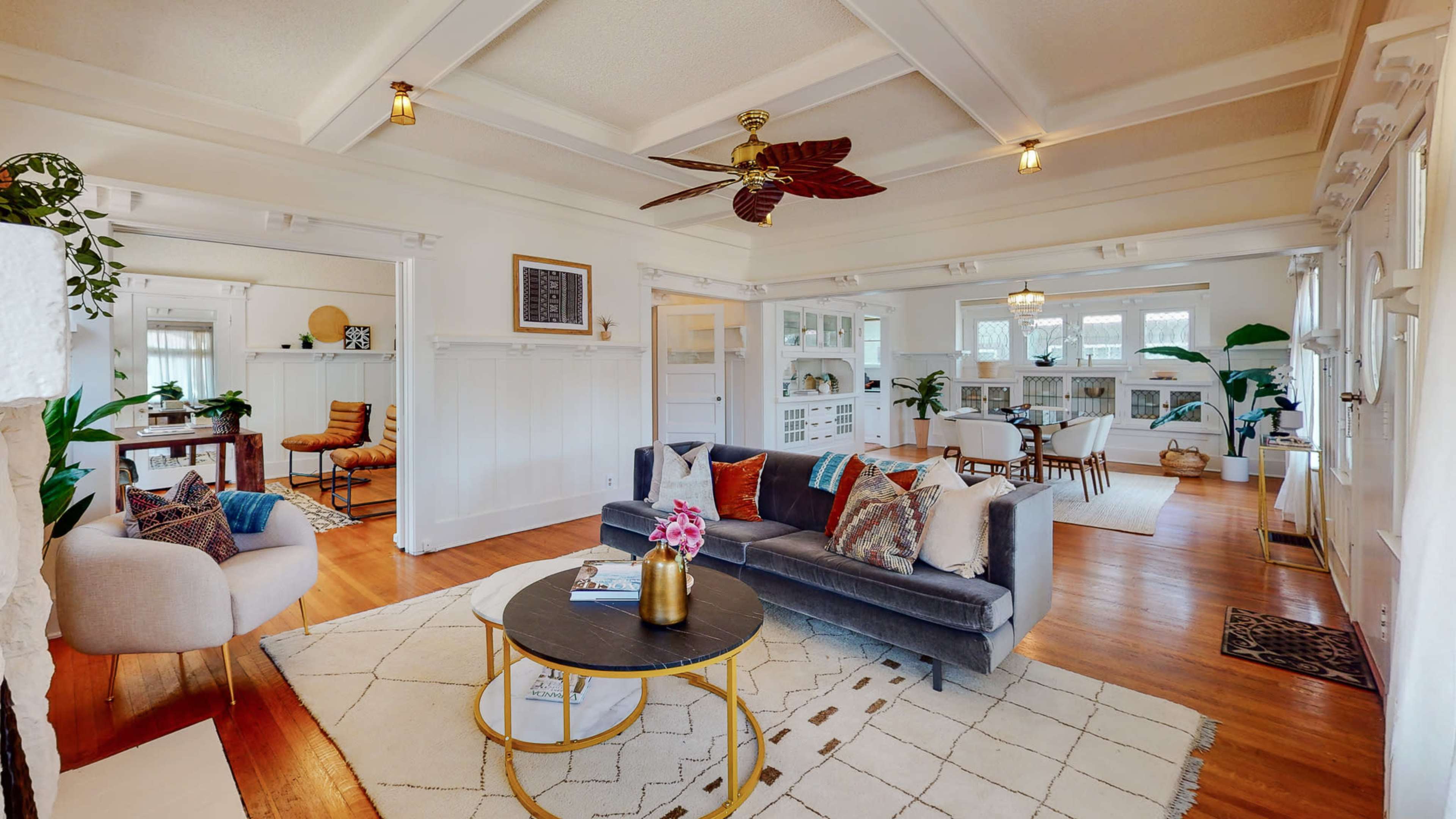 The image shows a spacious living area with a gray sofa, a round coffee table, and large windows, featuring a mix of indoor plants and wood flooring.