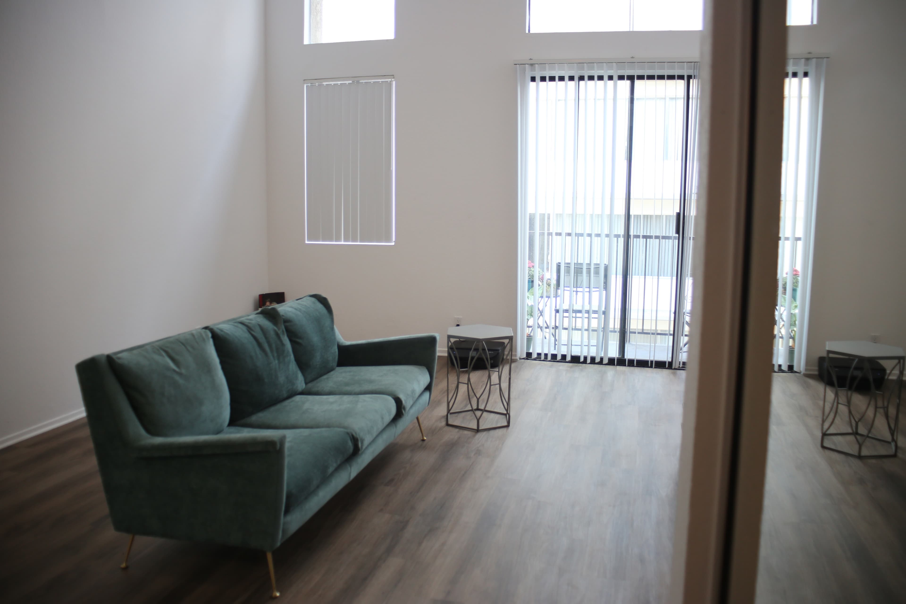 A minimalistic living room with a teal couch, two small side tables, and large windows with vertical blinds.