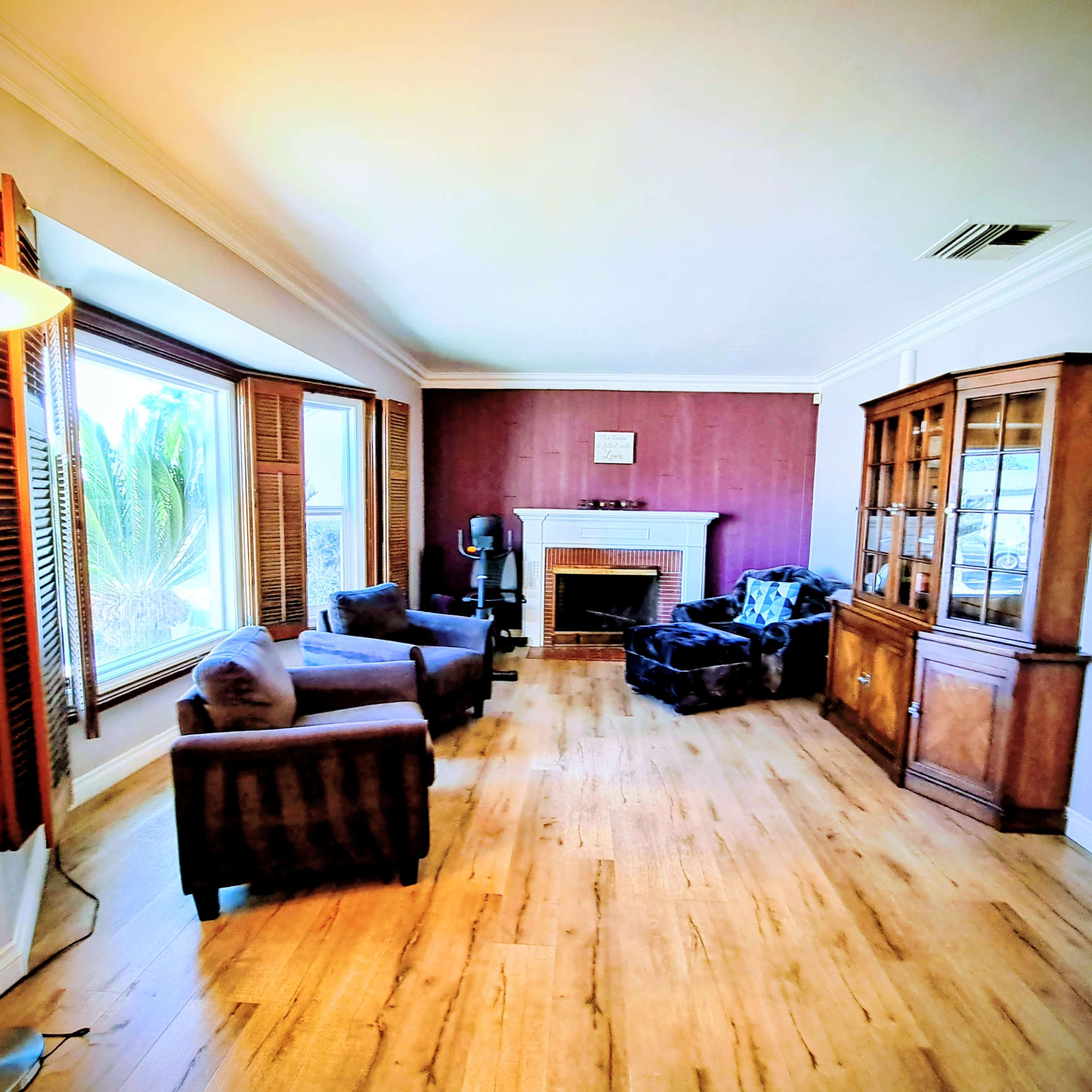 The image shows a cozy living room with two armchairs, a sofa, a fireplace, and a wooden cabinet, all arranged around a large window with plantation shutters.