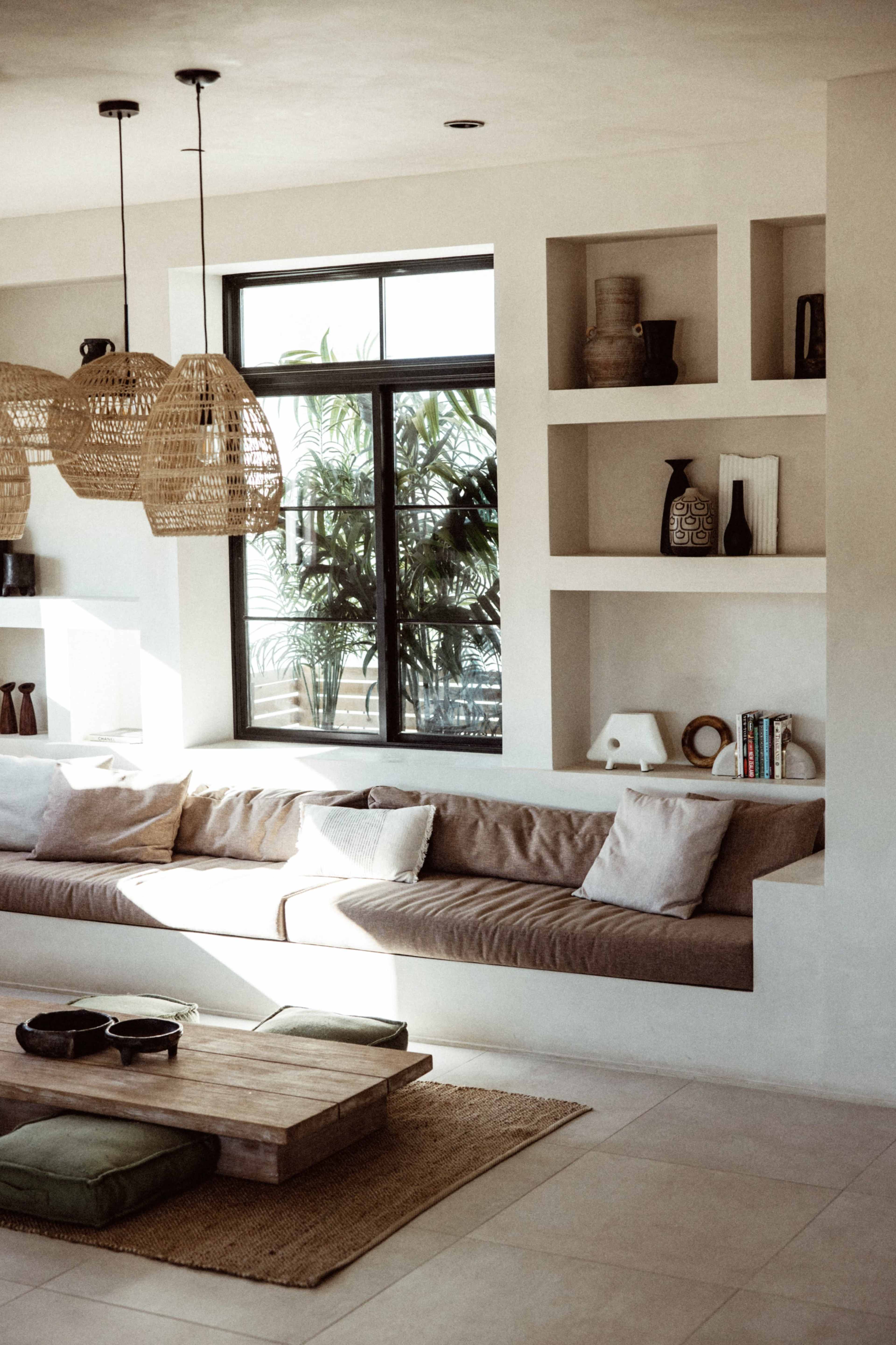 A modern living room with a long sofa against a wall of built-in shelves, filled with decorative items and plants, and a wooden coffee table on a light-colored floor.