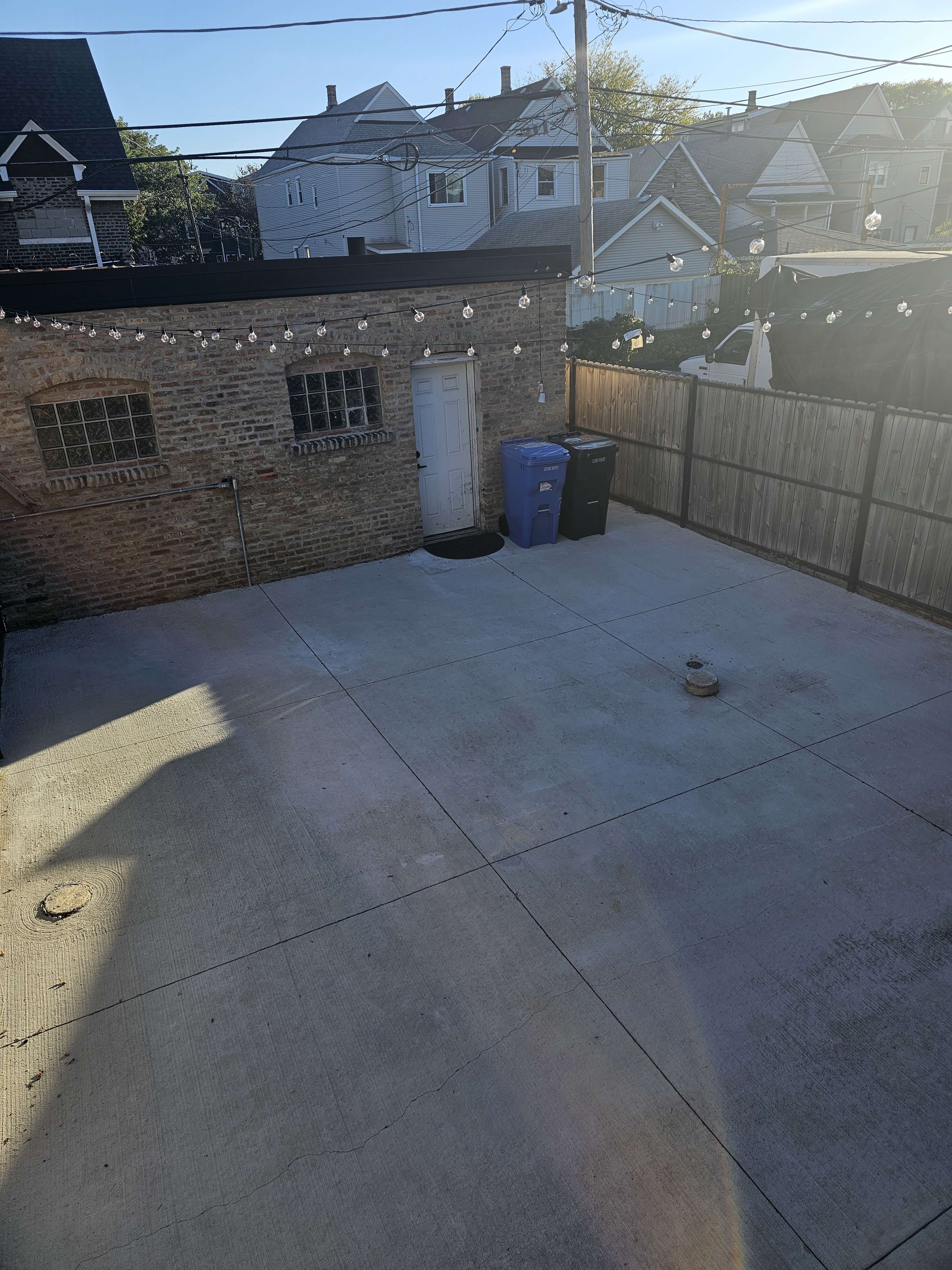 The image shows a clear, empty concrete backyard with a door, two trash bins, and a wooden fence in the background.