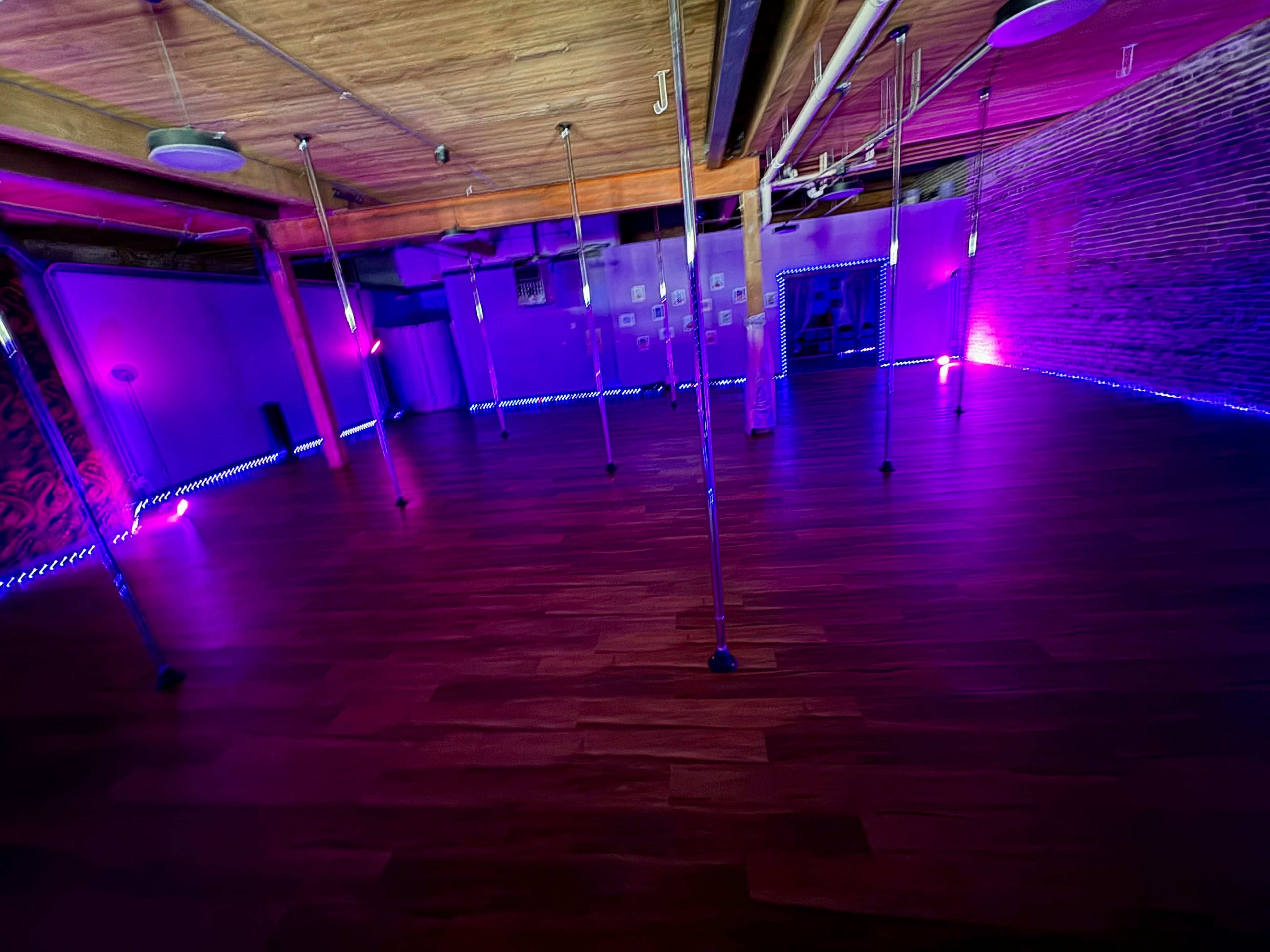 The image shows a dance studio with polished wooden floors, multiple poles, and purple lighting along the walls and floor.