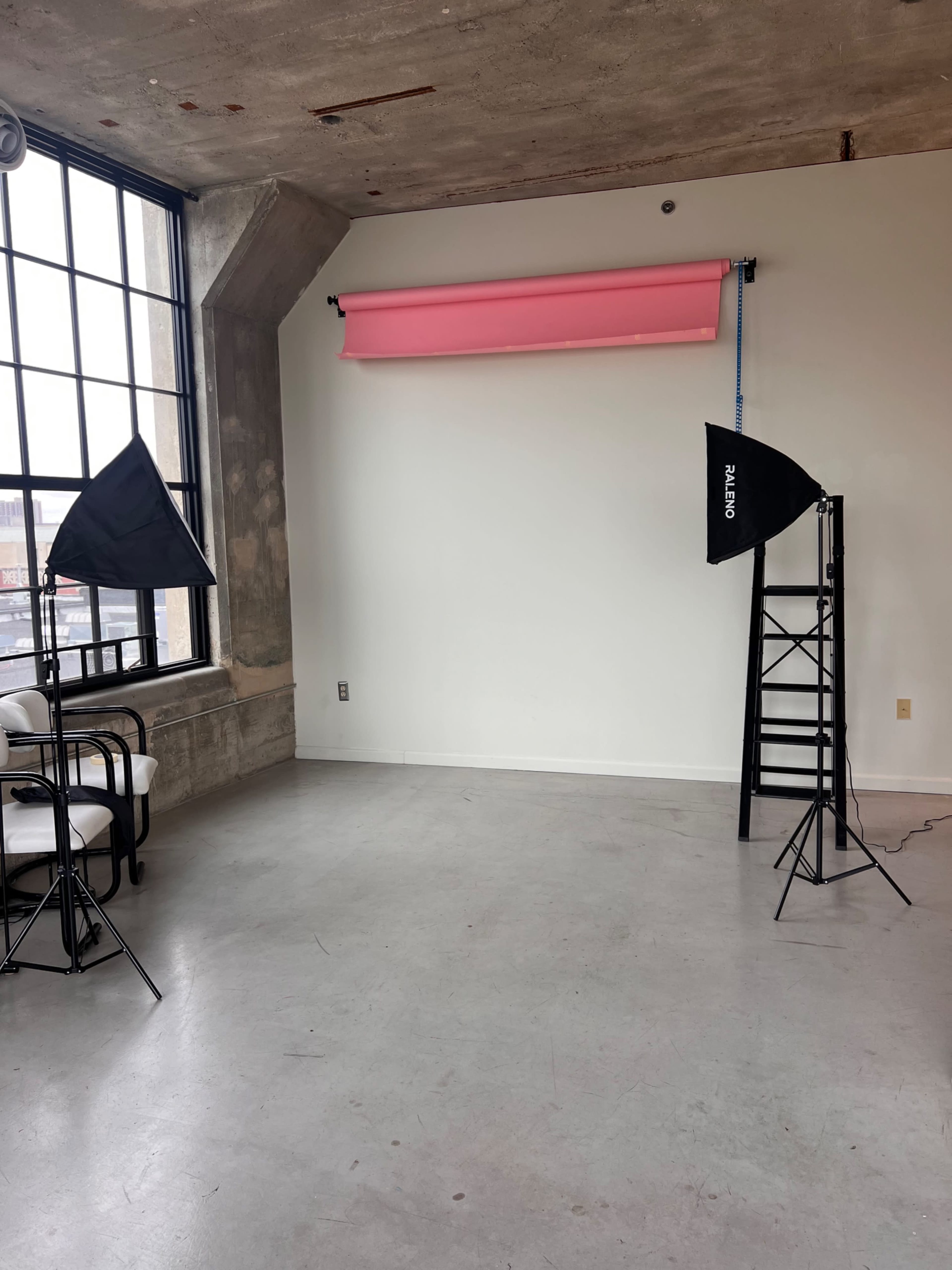 The image shows a photography studio with concrete walls, large windows, and two light stands positioned on either side of a blank wall featuring a pink backdrop.