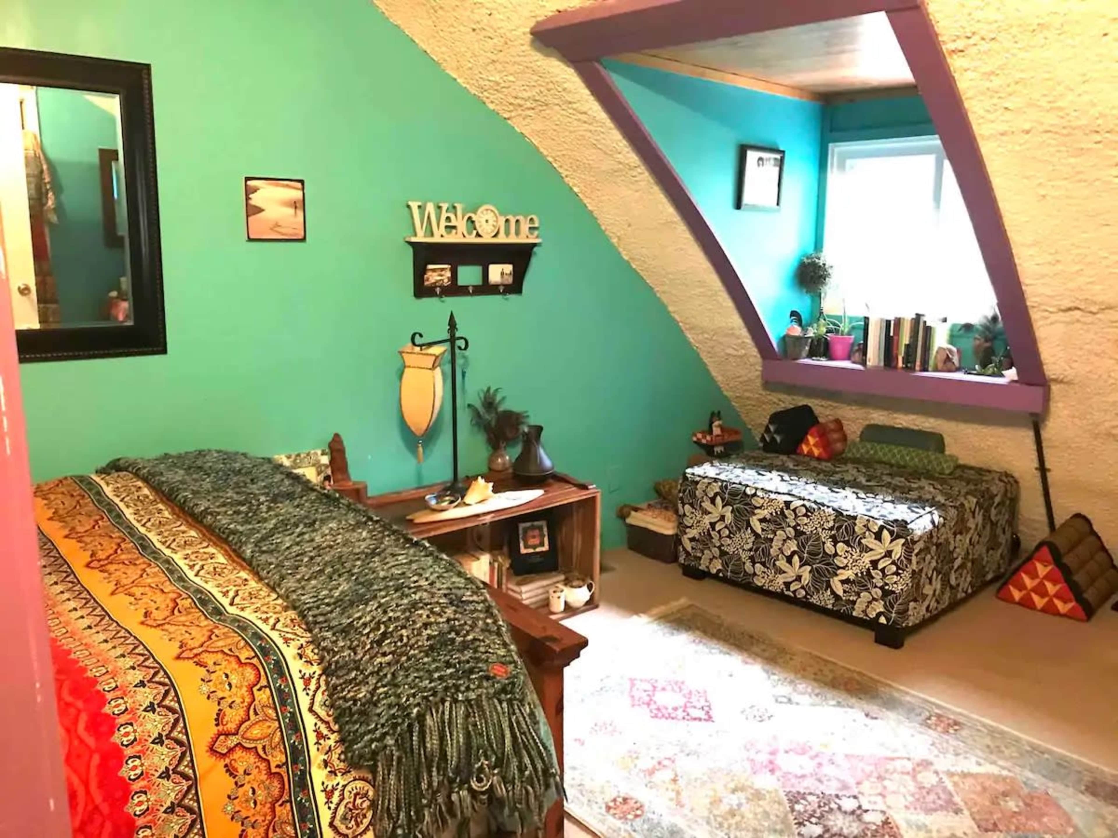 The image shows a cozy, colorful bedroom with an arched ceiling, a bed with a patterned throw, a small seating area, and decorative wall art.