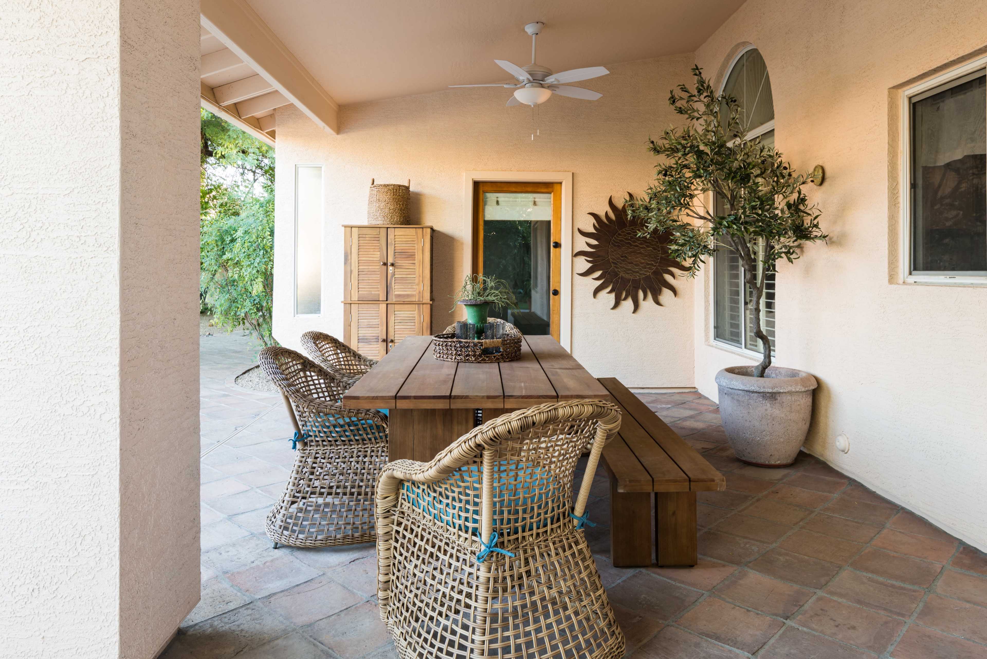 A covered patio features a wooden dining table surrounded by woven chairs, with a potted plant and decorative wall piece nearby.