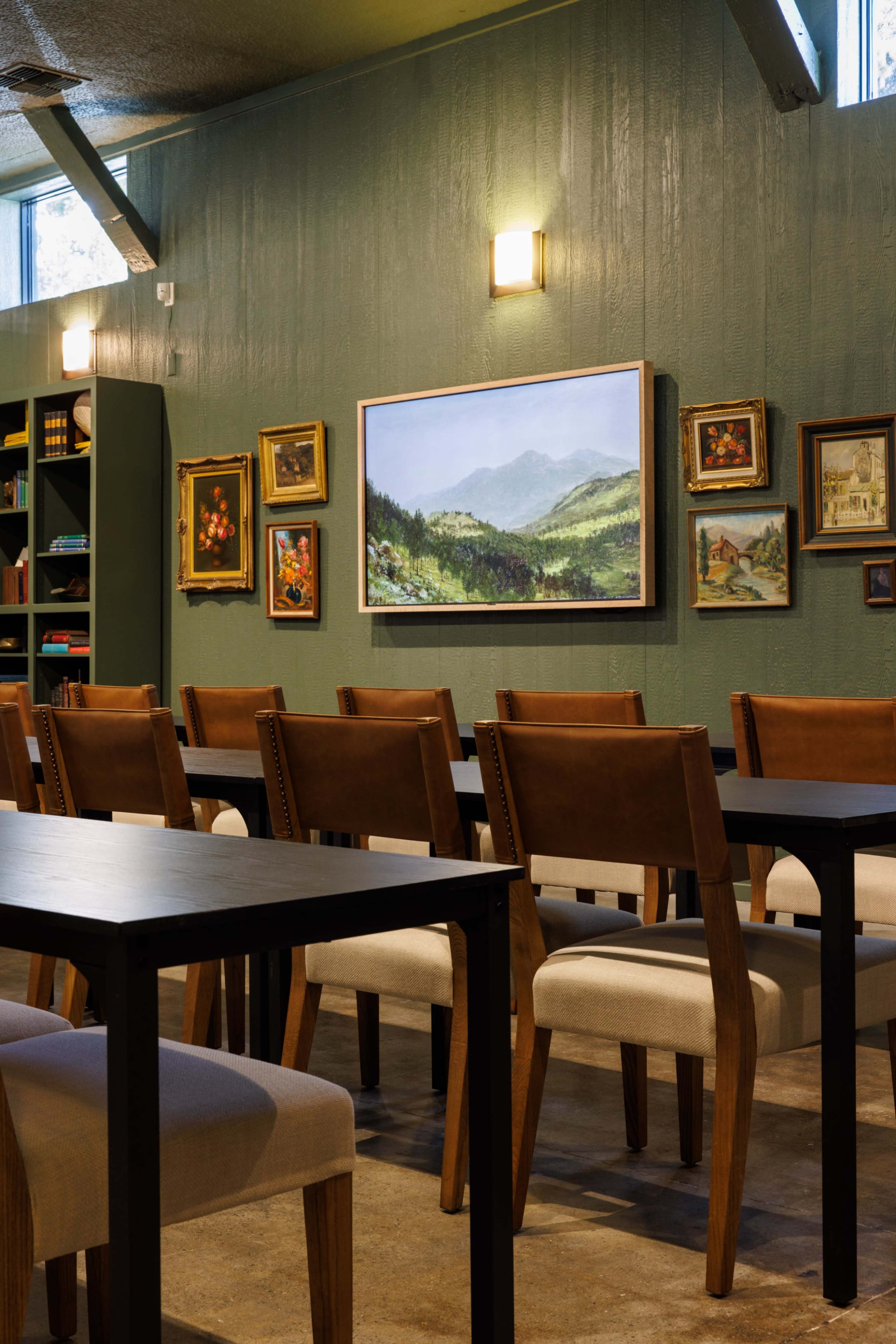 The image shows a dining area with wooden tables and chairs, featuring a green wall adorned with various framed artworks.
