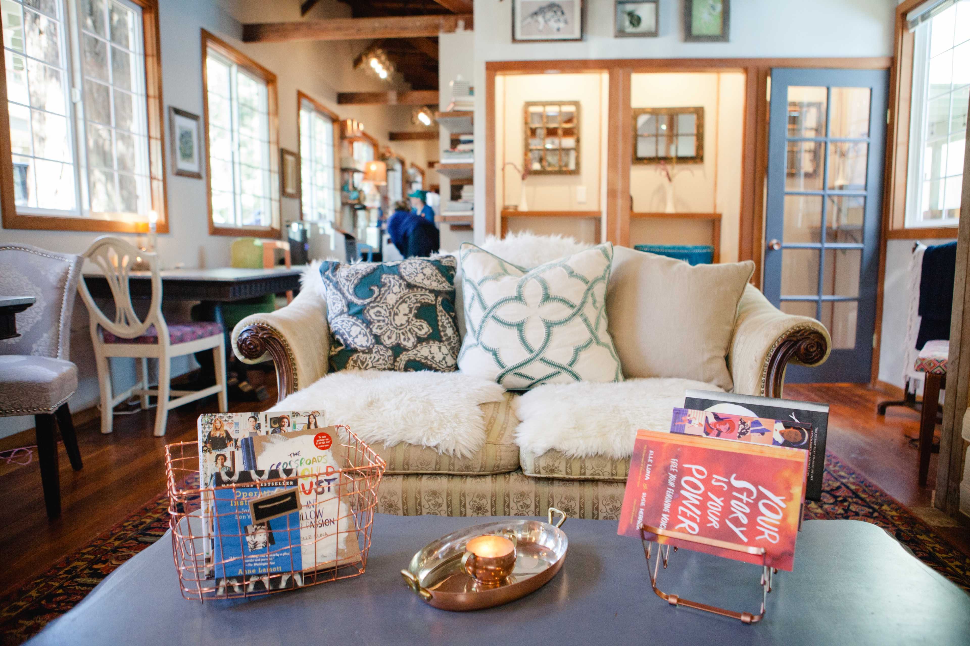 A cozy living room with a patterned sofa, a coffee table displaying books and a candle, and large windows allowing natural light to illuminate the space.