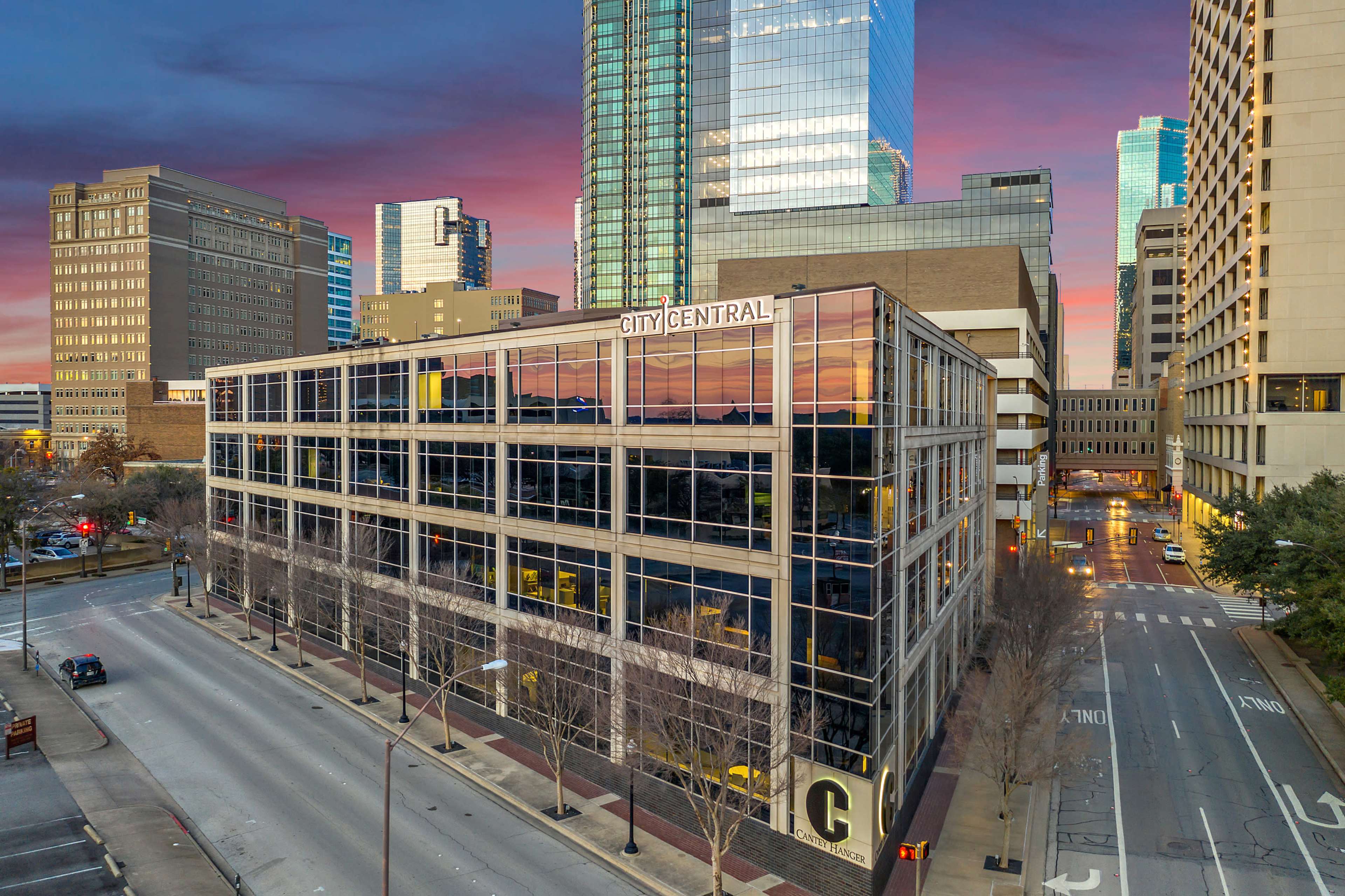 The image shows a modern office building with reflective glass windows, located at the corner of a city street, under a colorful sky at sunset.