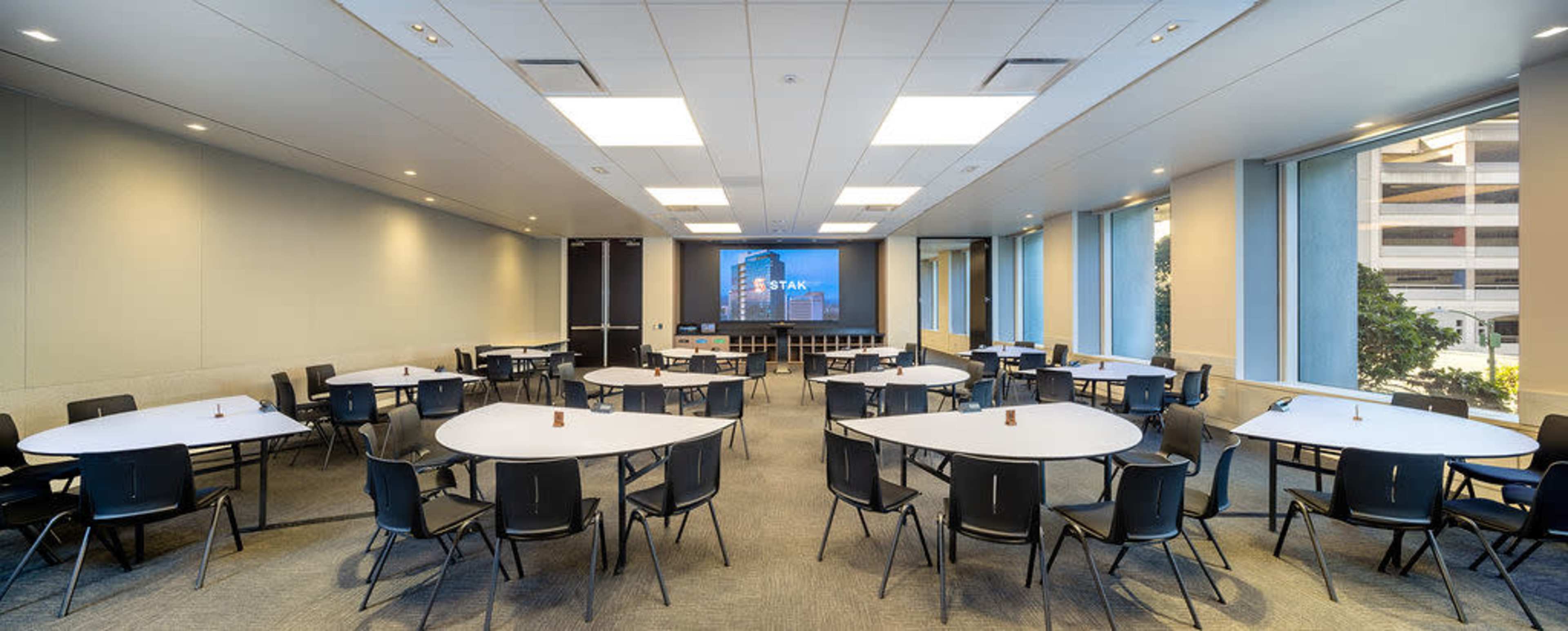 The image shows a modern conference room arranged with multiple round tables and chairs, featuring large windows and a projection screen at the front.