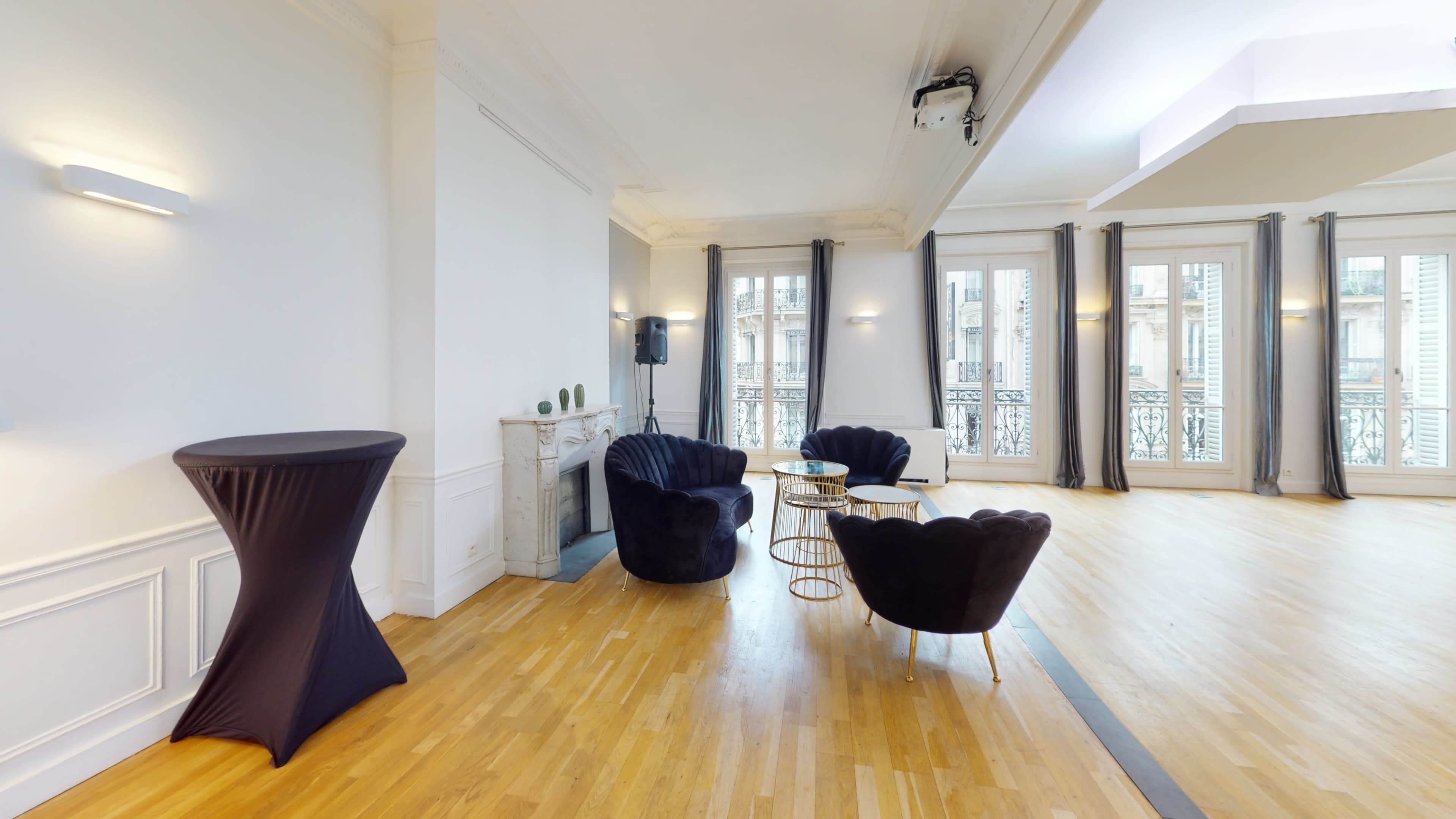 The image shows an elegant room with parquet flooring, featuring two black armchairs, a round table, a tall cocktail table, and large windows overlooking an exterior balcony.