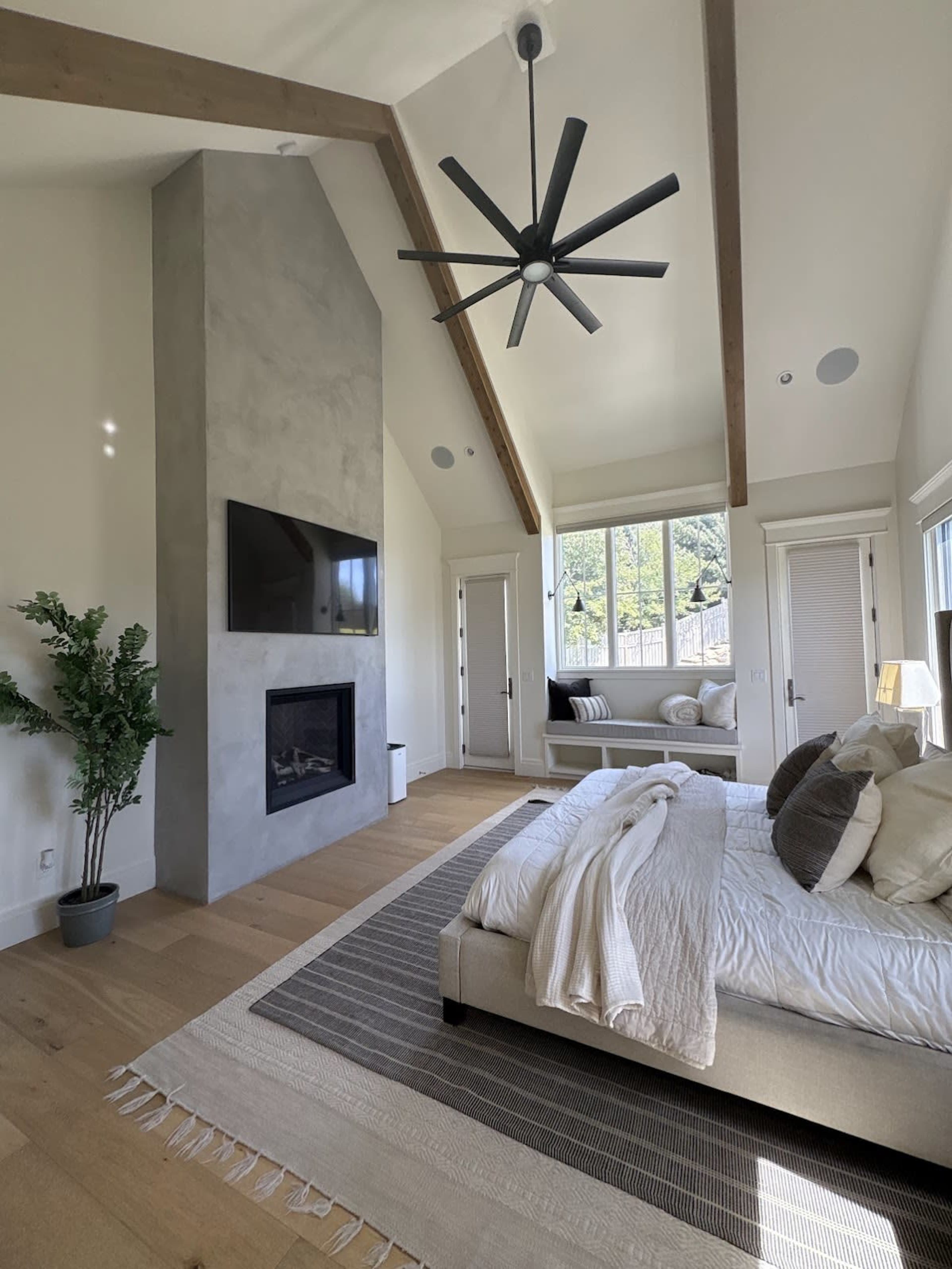 The image shows a spacious bedroom featuring a high ceiling with wooden beams, a large flat-screen TV mounted on a concrete wall, and a bed with white linens positioned on a hardwood floor.