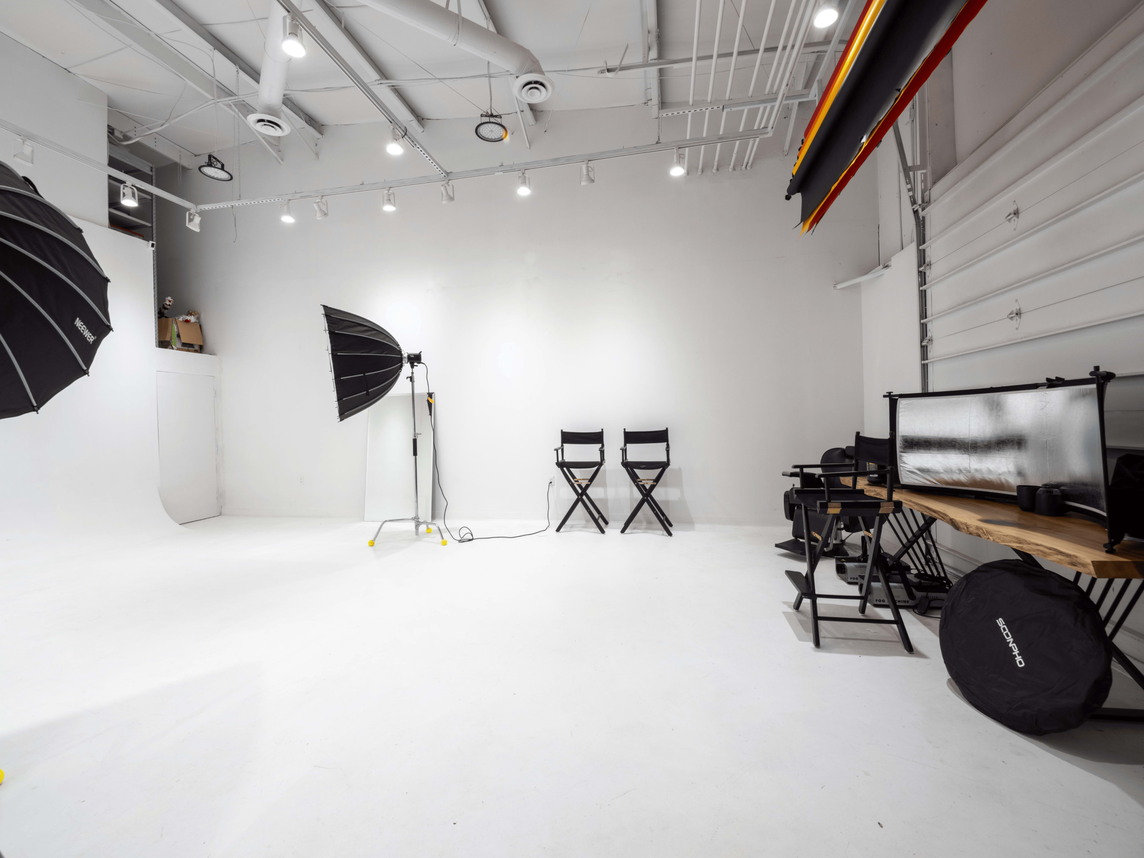 An empty photography studio with white walls, a wooden table, and various lighting equipment.