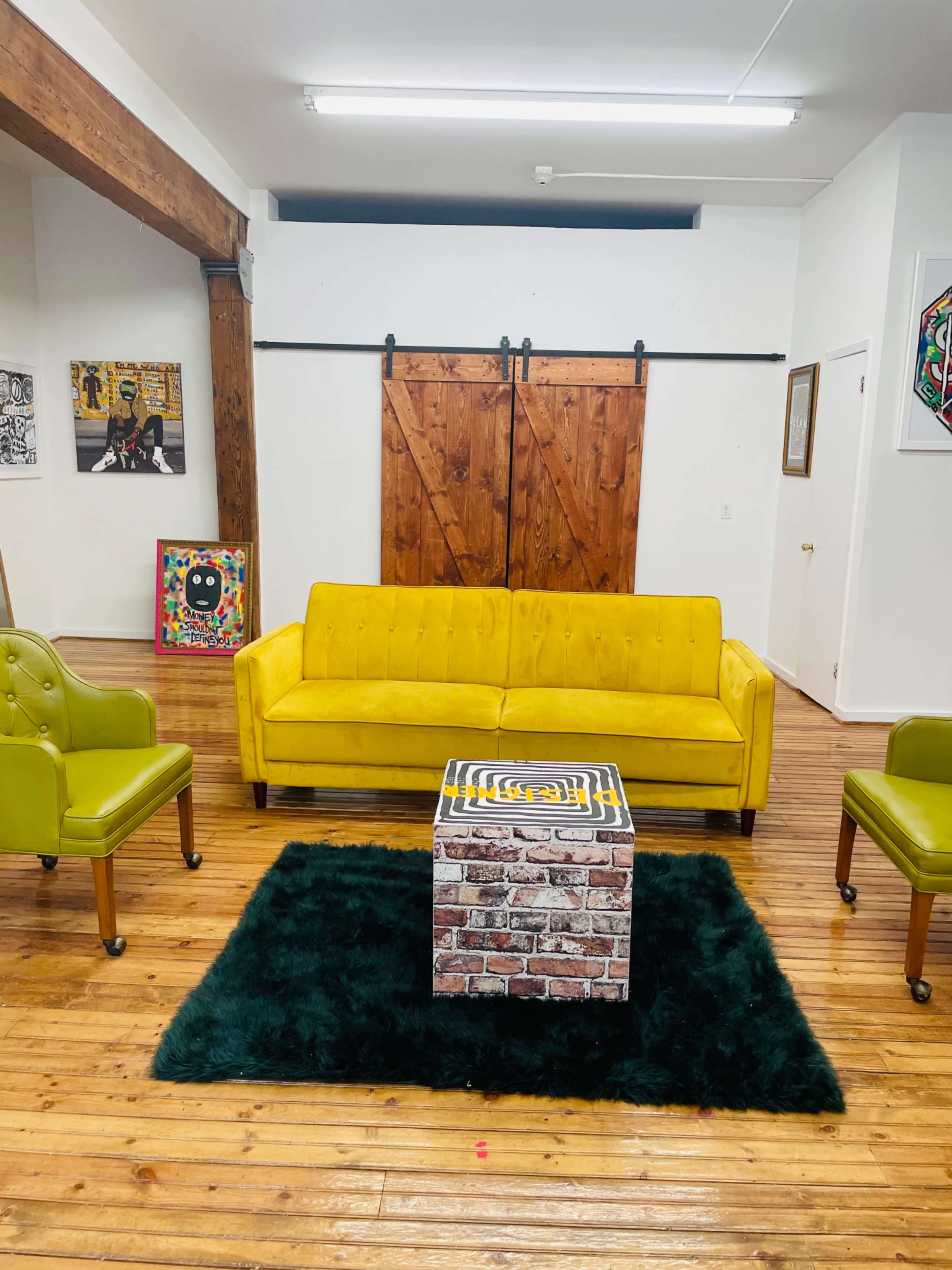 A yellow sofa, two green armchairs, and a square coffee table on a dark rug in a wooden-floored interior space.