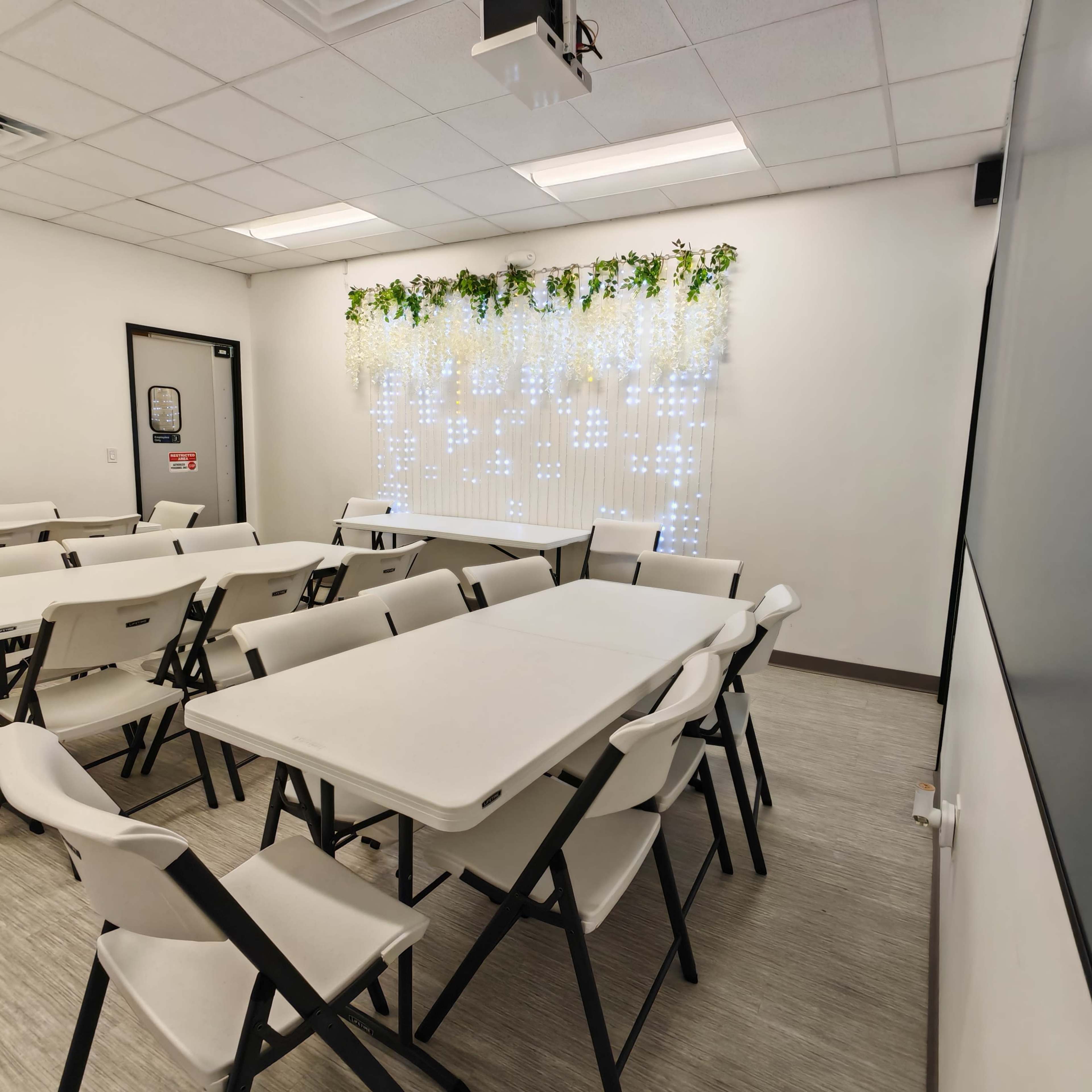 A classroom features several folding tables arranged in a U-shape, with white chairs and a backdrop of decorative lights and plants on a wall.