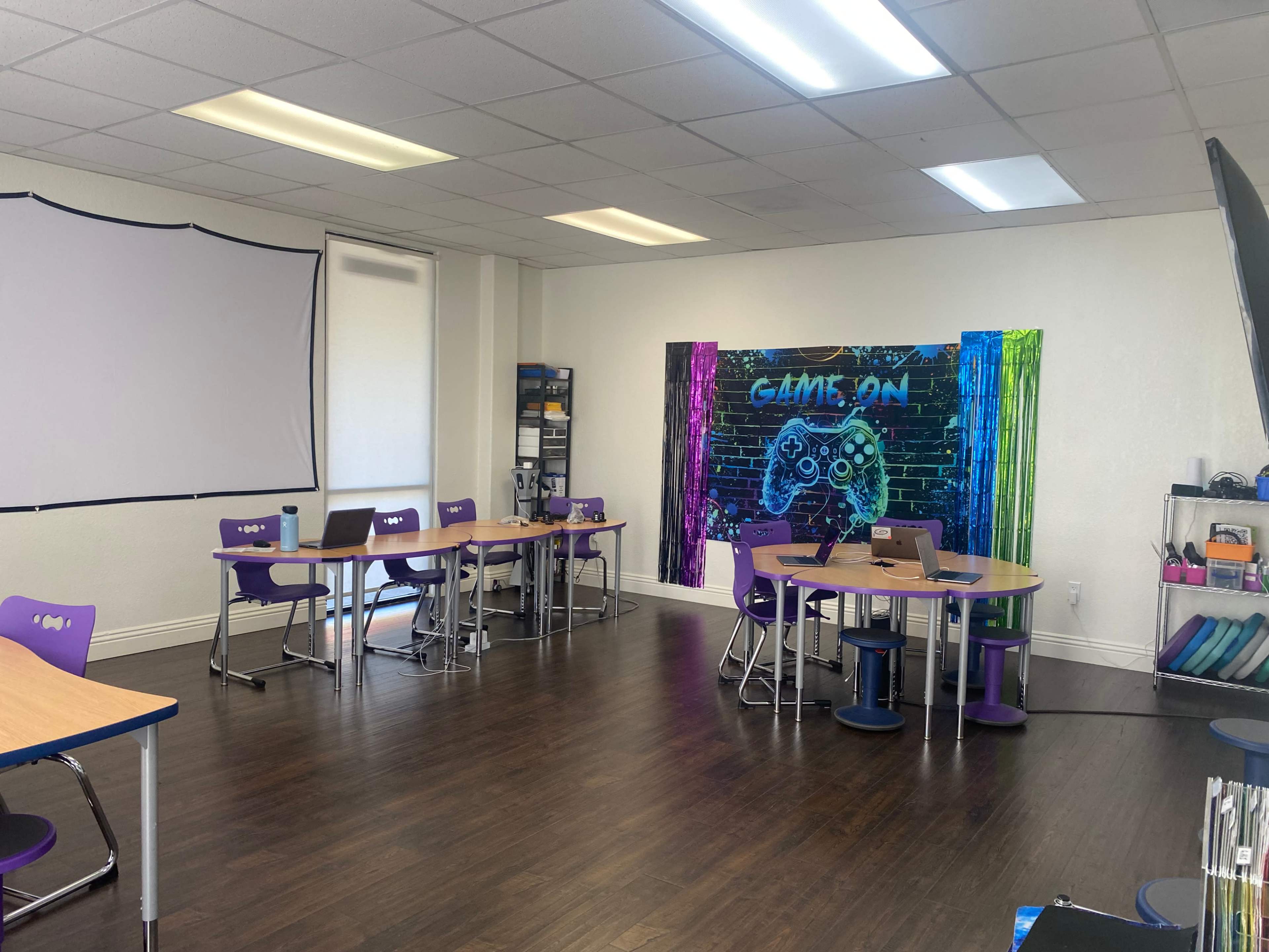 The image shows a classroom with various tables and chairs arranged, a projector screen, and a colorful wall mural featuring a gaming theme.