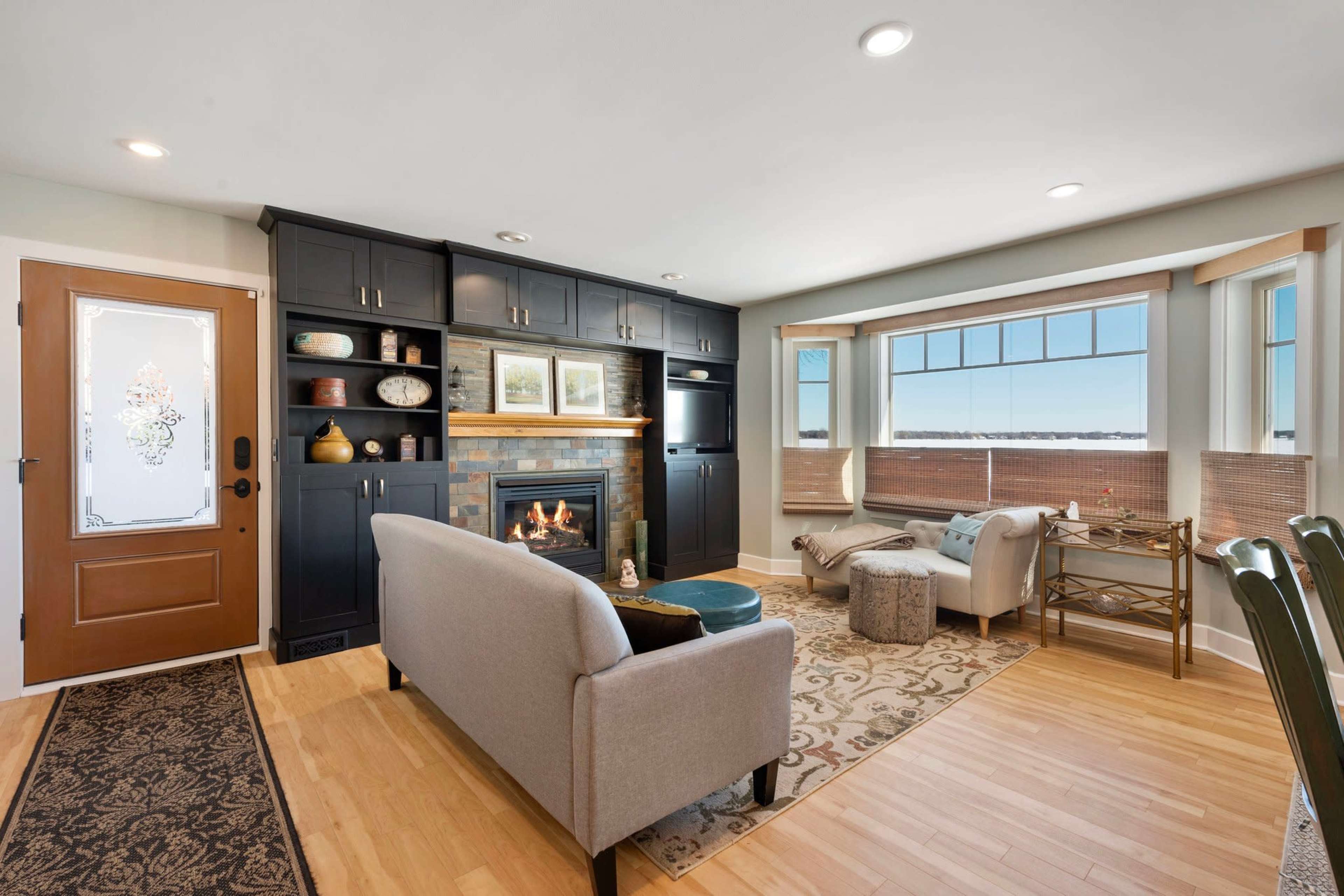 A cozy living room features a fireplace, natural light from large windows, and modern furnishings including a gray sofa and a decorative armchair.
