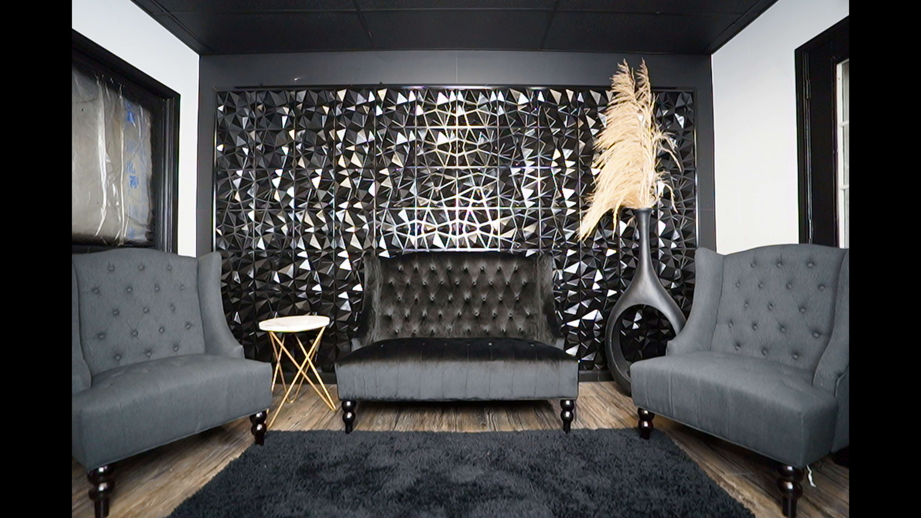 The image shows a modern living space with two gray armchairs flanking a black tufted couch, set against a geometric patterned wall and a decorative plant.