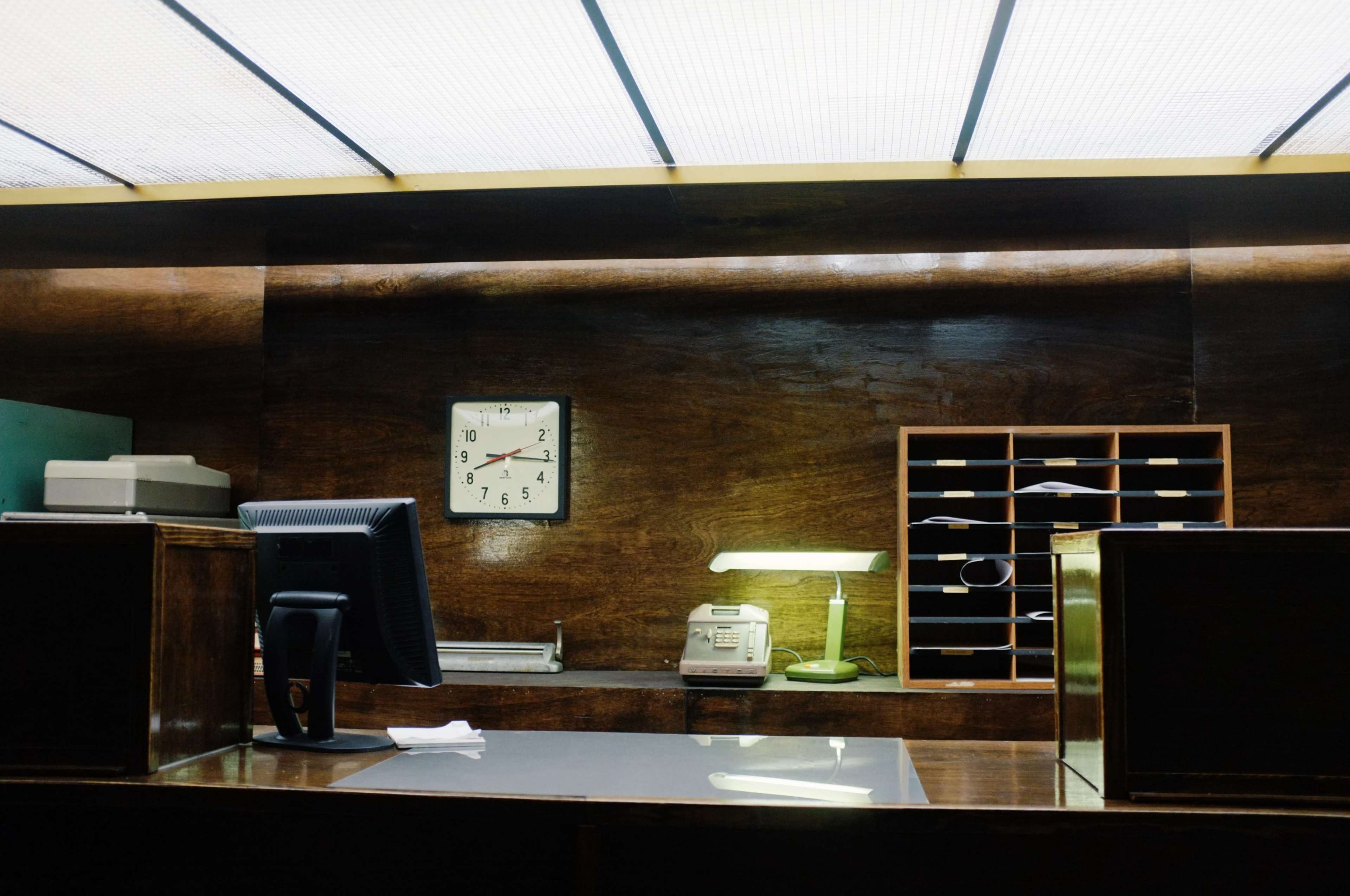 A wooden reception desk with a clock, computer monitor, phone, and an organized mail slot in a dimly lit office.