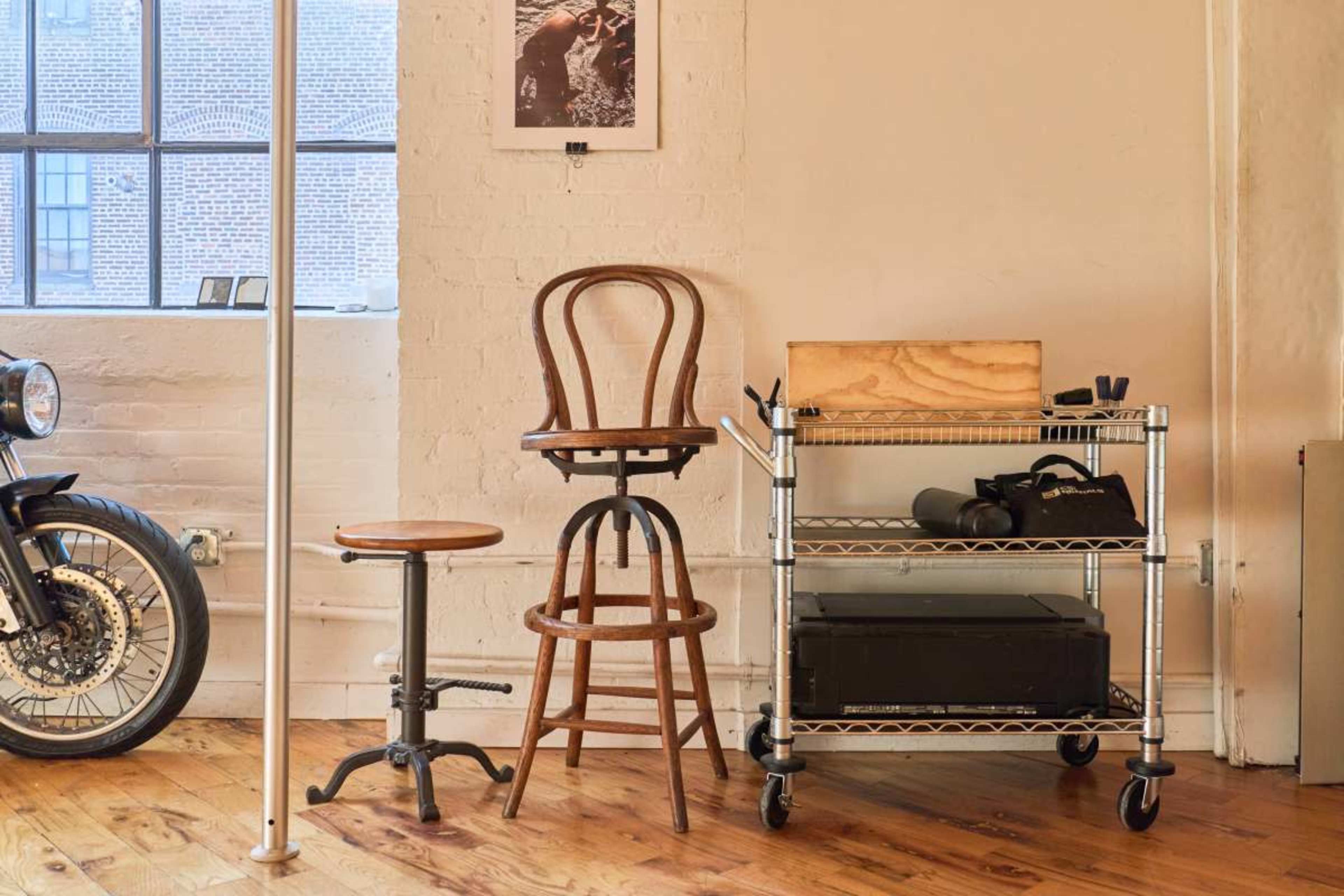 The image shows a workspace with a wooden stool, a tall wooden barstool, and a metal cart holding various items, alongside a motorcycle and a window.