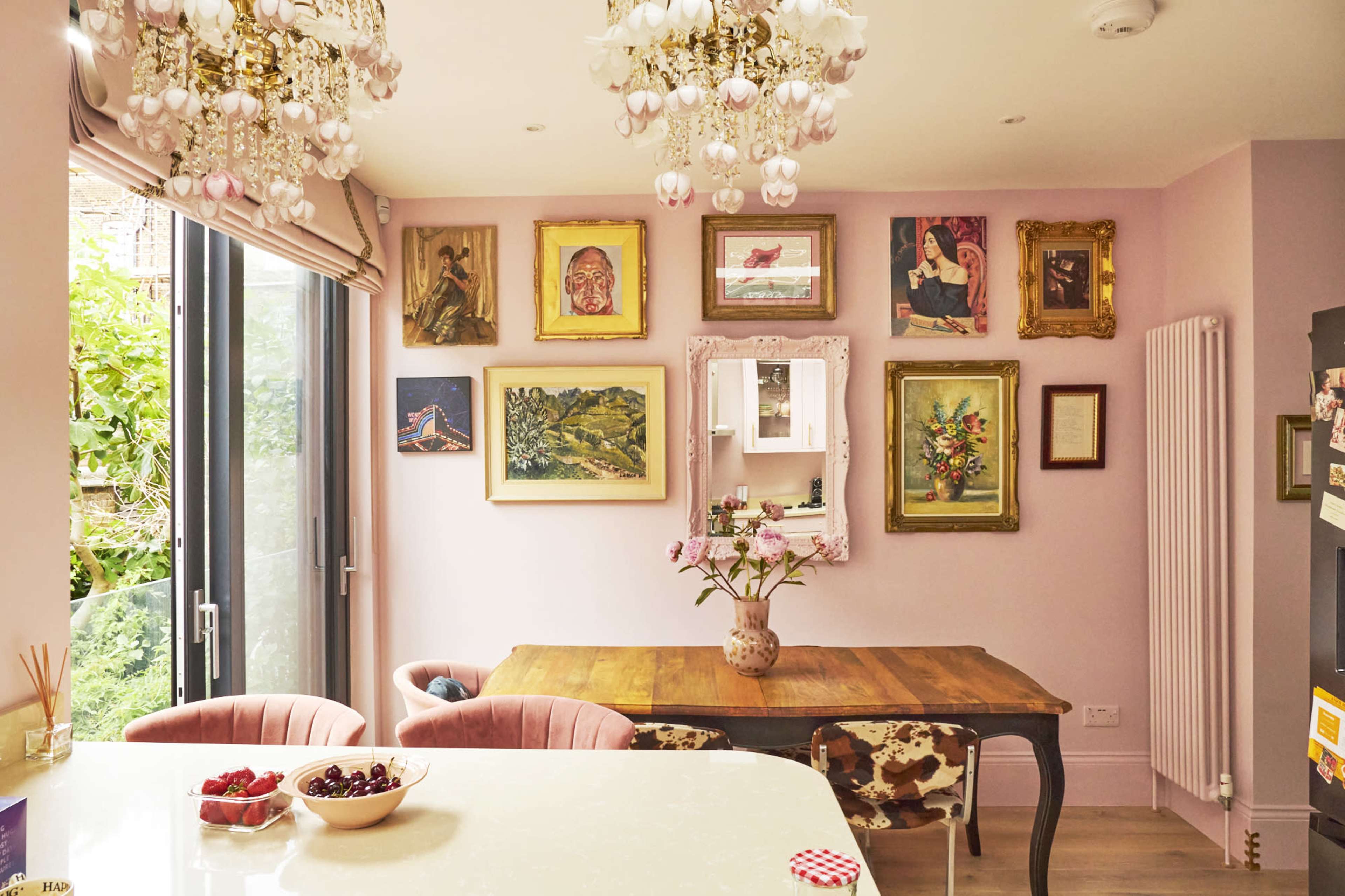 A dining area with a wooden table, pink chairs, and walls adorned with various framed artworks, illuminated by decorative chandeliers.