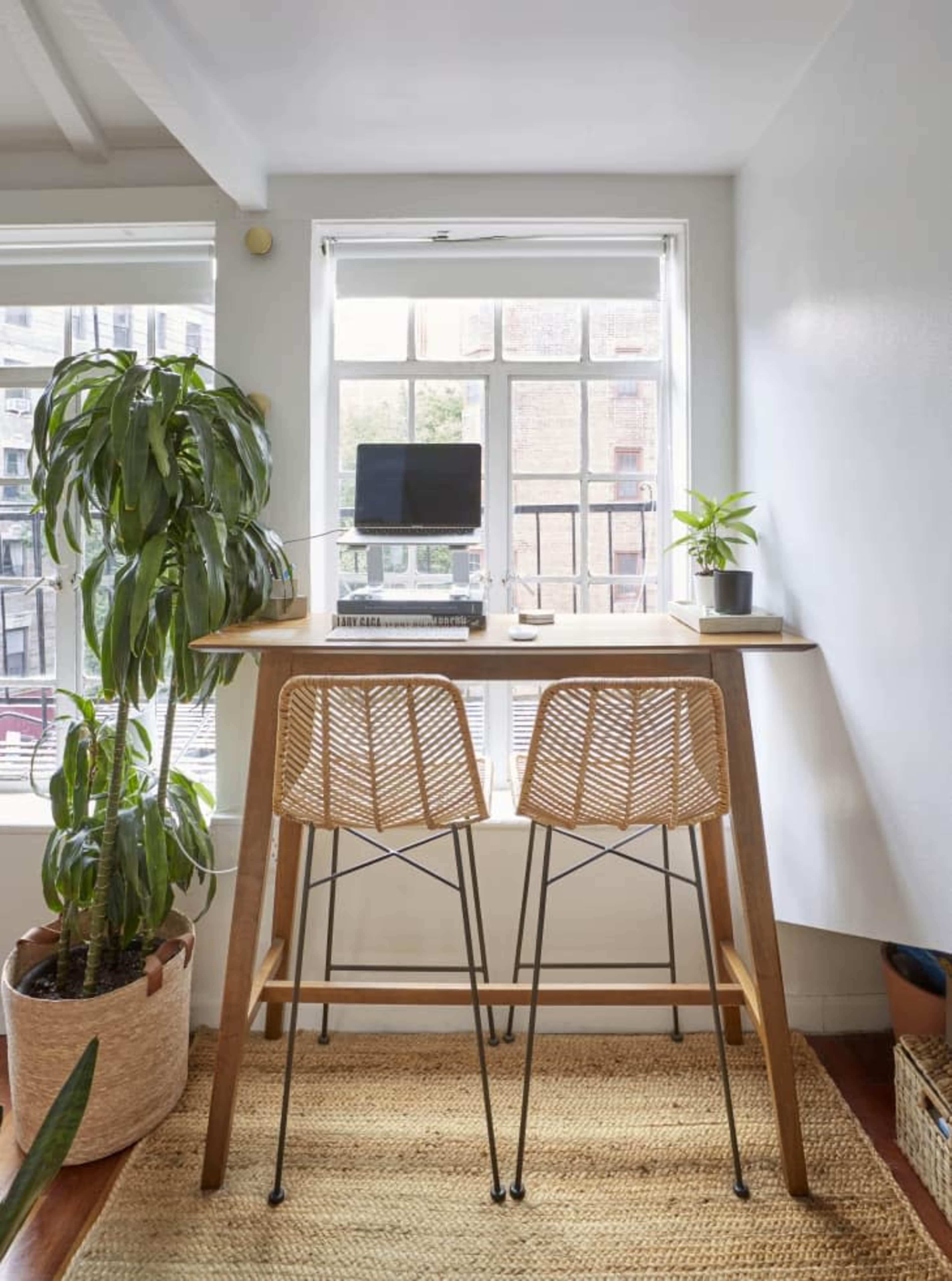A small workspace features a wooden table with two woven chairs, positioned near a window with a potted plant and a computer on top.