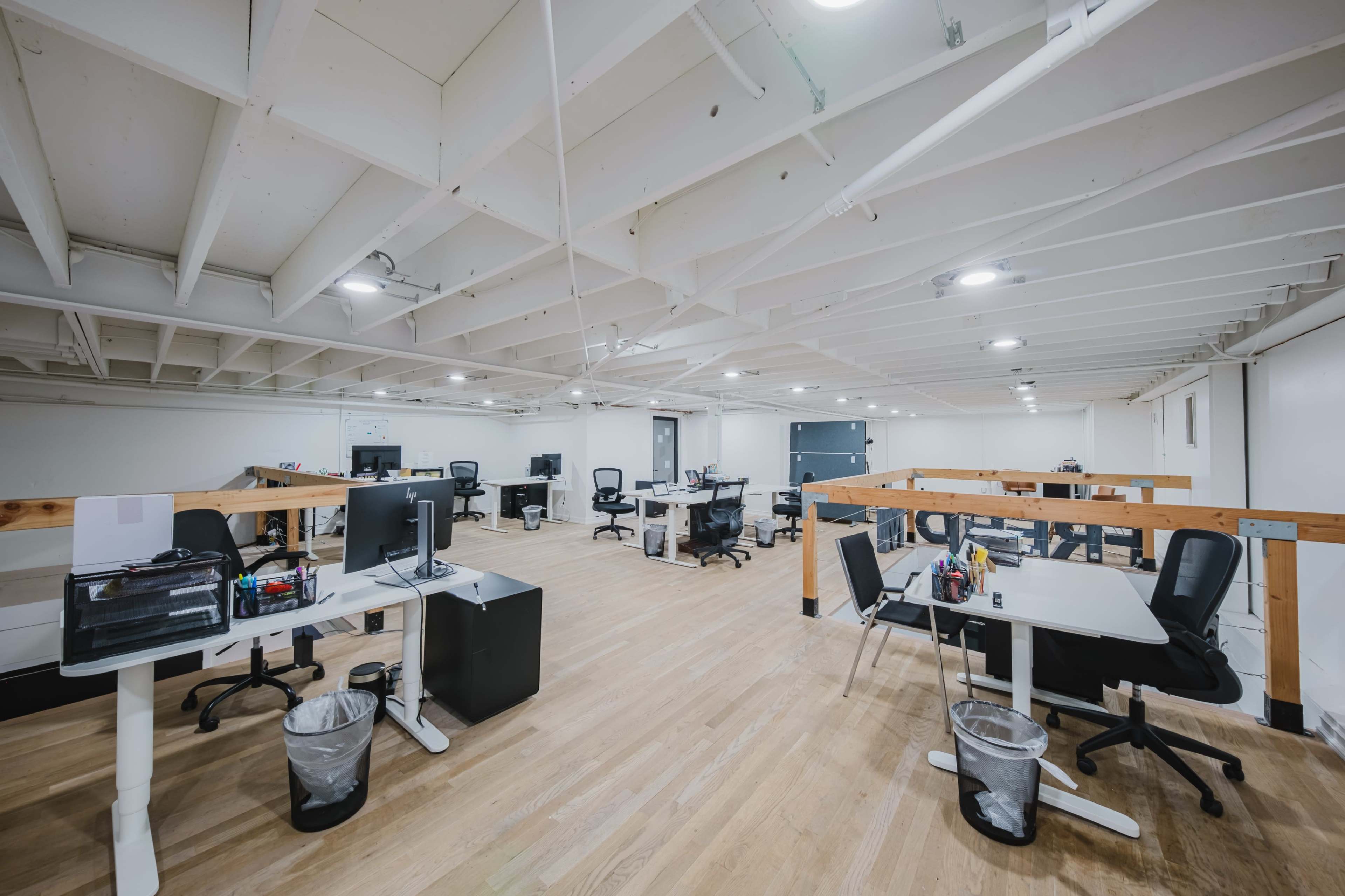 The image shows a modern office space with several workstations, including desks and chairs, arranged under a high ceiling.