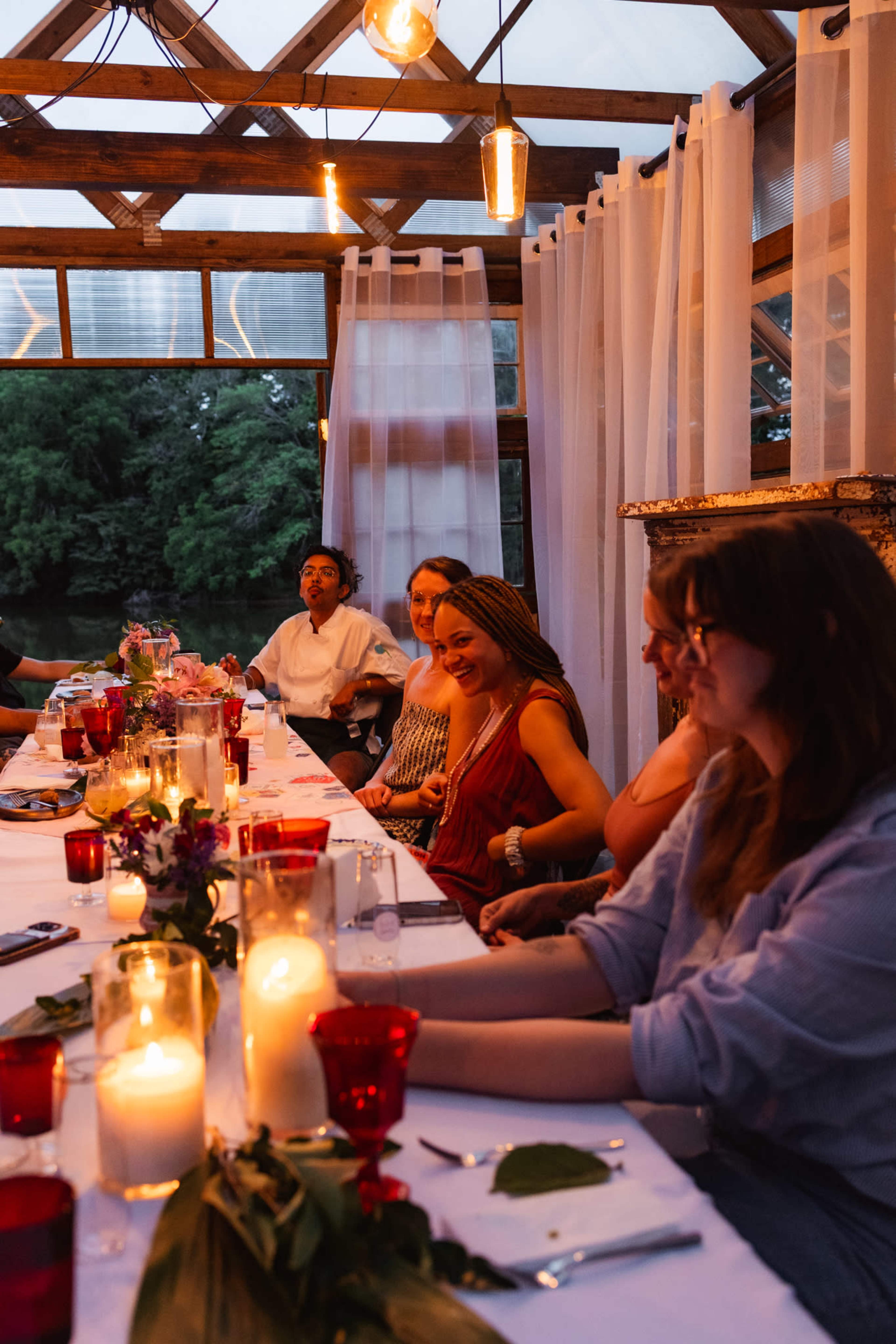 A group of people are seated at a long dinner table adorned with candles and flowers, enjoying a meal in a softly lit, rustic setting.