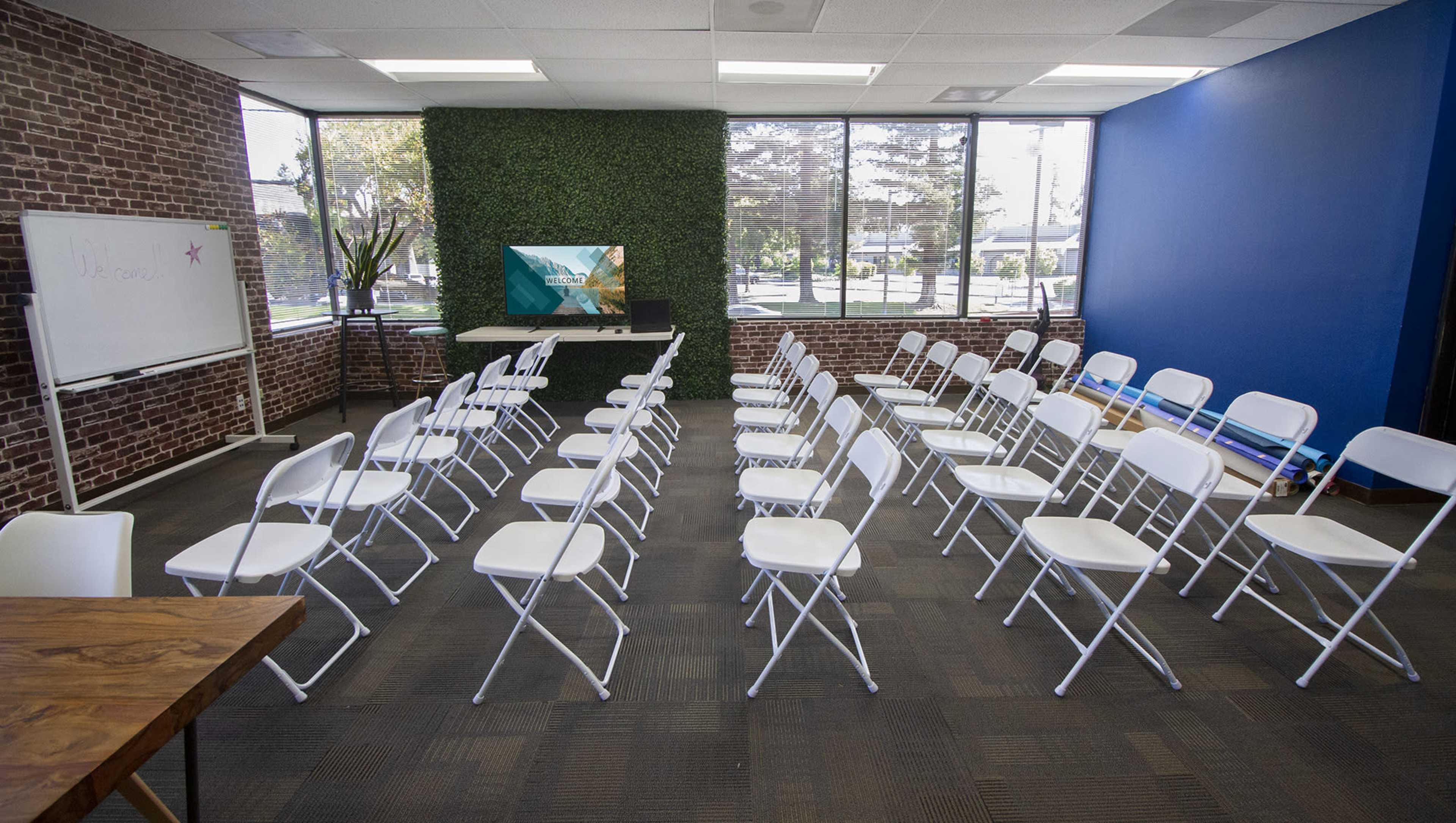 A conference room features rows of white folding chairs facing a television screen on a table, with brick walls and large windows providing natural light.