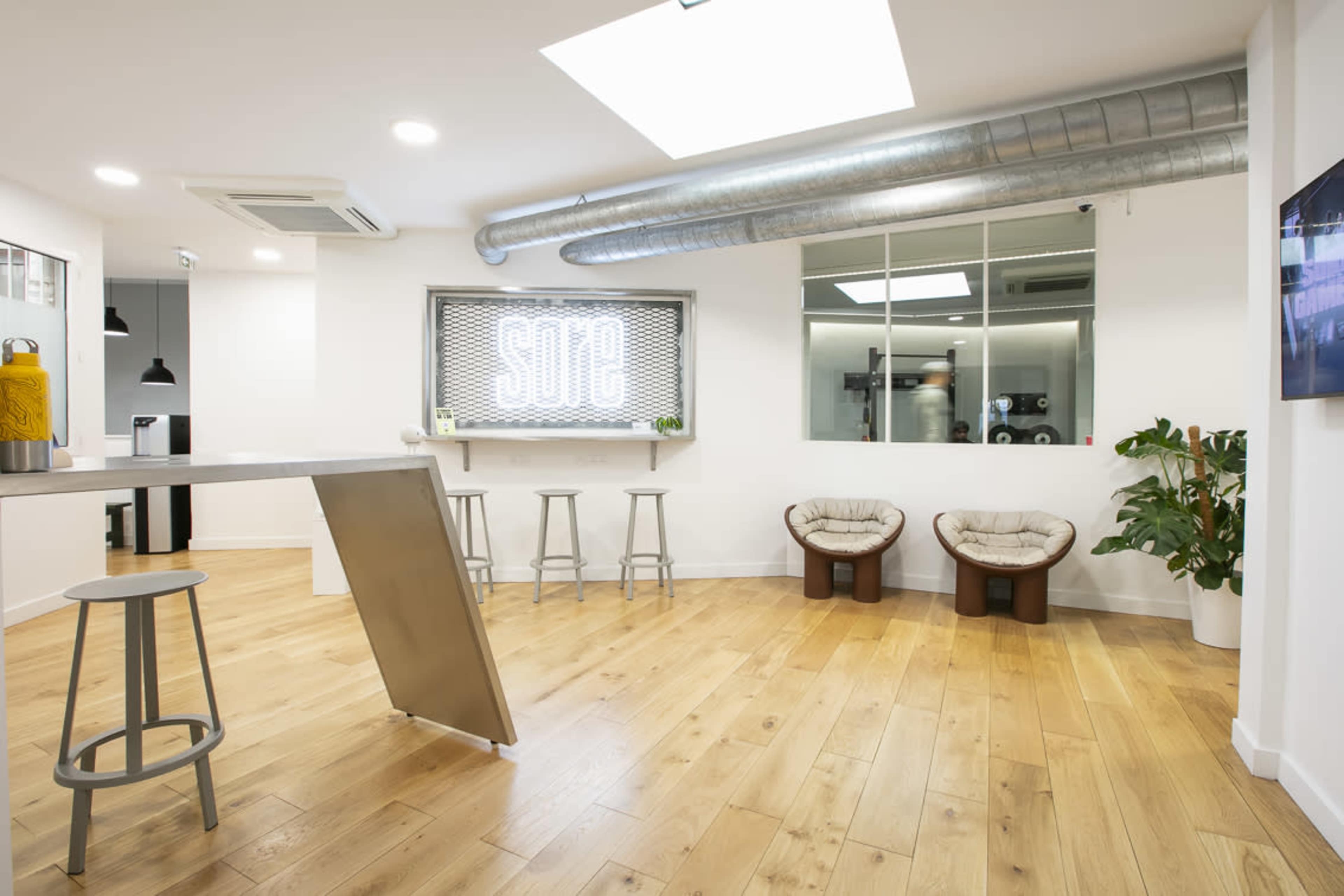 A modern office space features wooden flooring, a metal table with stools, and two plush chairs next to a large window displaying the word "SODE."