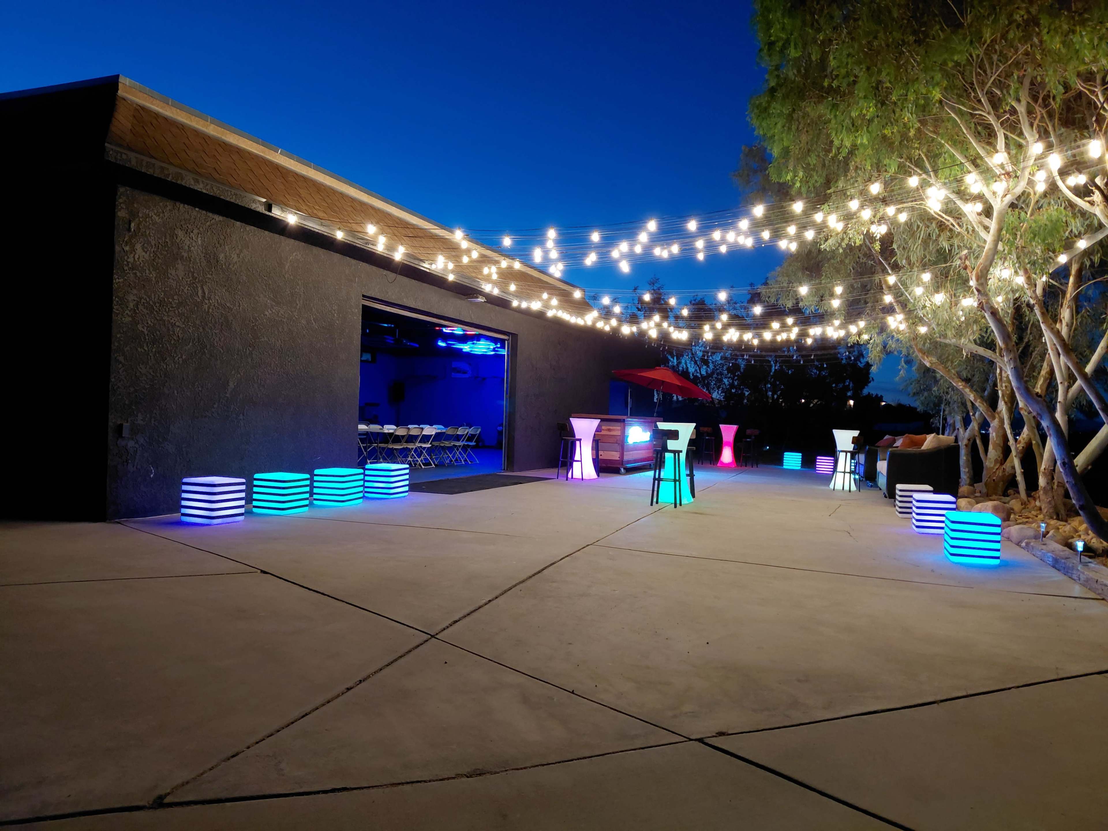 The scene shows a dimly lit outdoor area with decorative string lights, illuminated furniture in various colors, and a spacious concrete floor adjacent to a building.