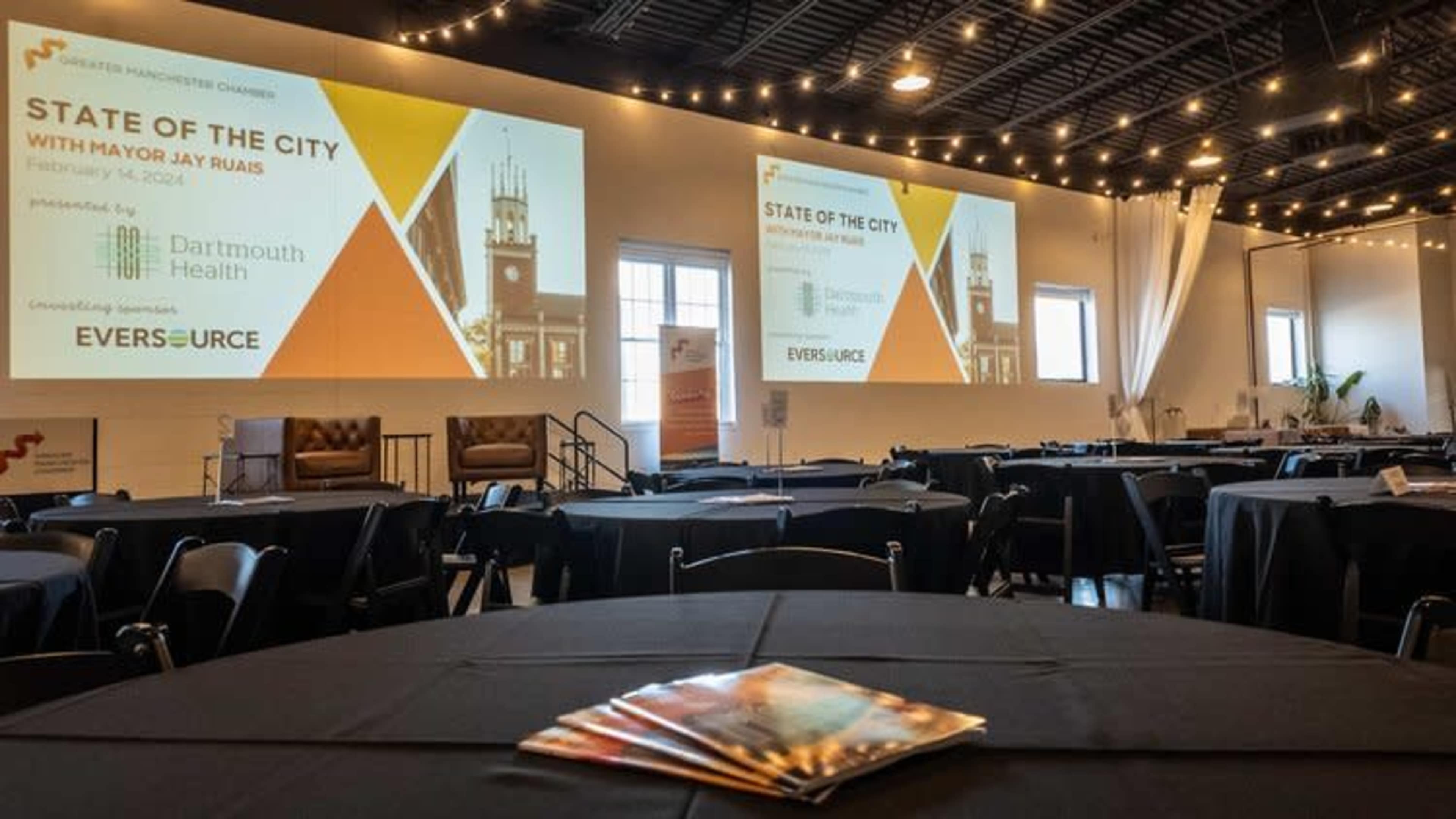 An event space is set up with round tables and a large screen displaying the "State of the City" presentation featuring Mayor Jay Rojas.