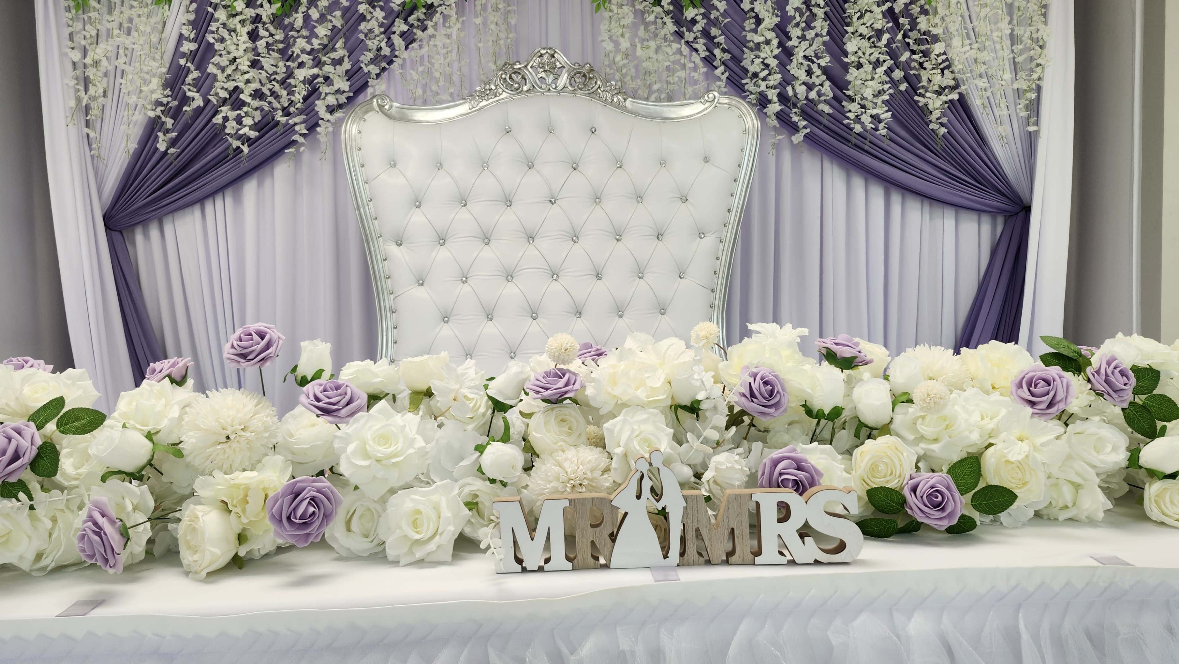 A decorated wedding table features a large white chair with a backdrop of purple drapery and an arrangement of white and purple flowers, along with a "Mr & Mrs" sign.