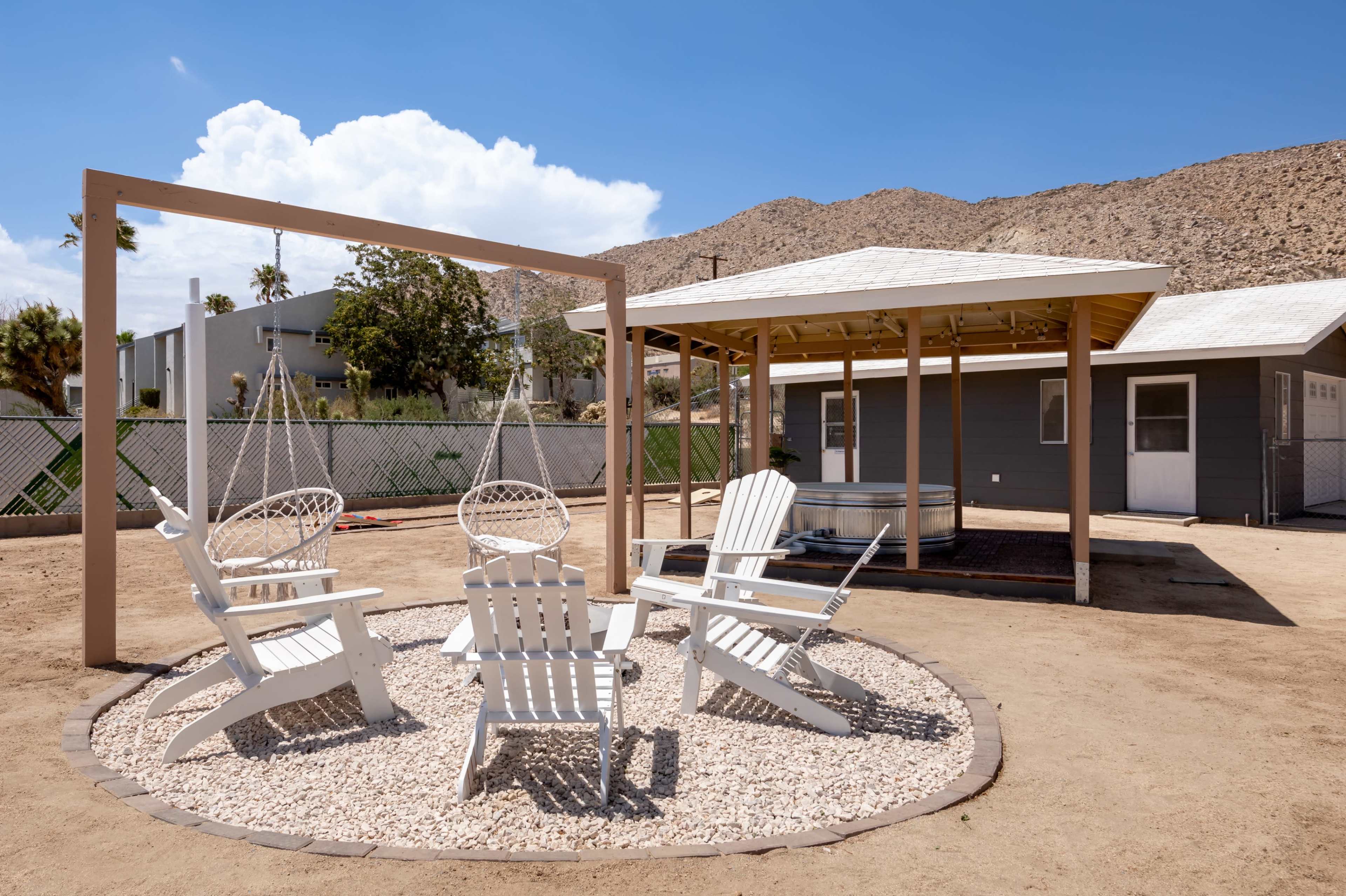 The image shows a sandy outdoor area with white patio chairs arranged in a circle around a gravel base, next to a gazebo and surrounded by mountains.