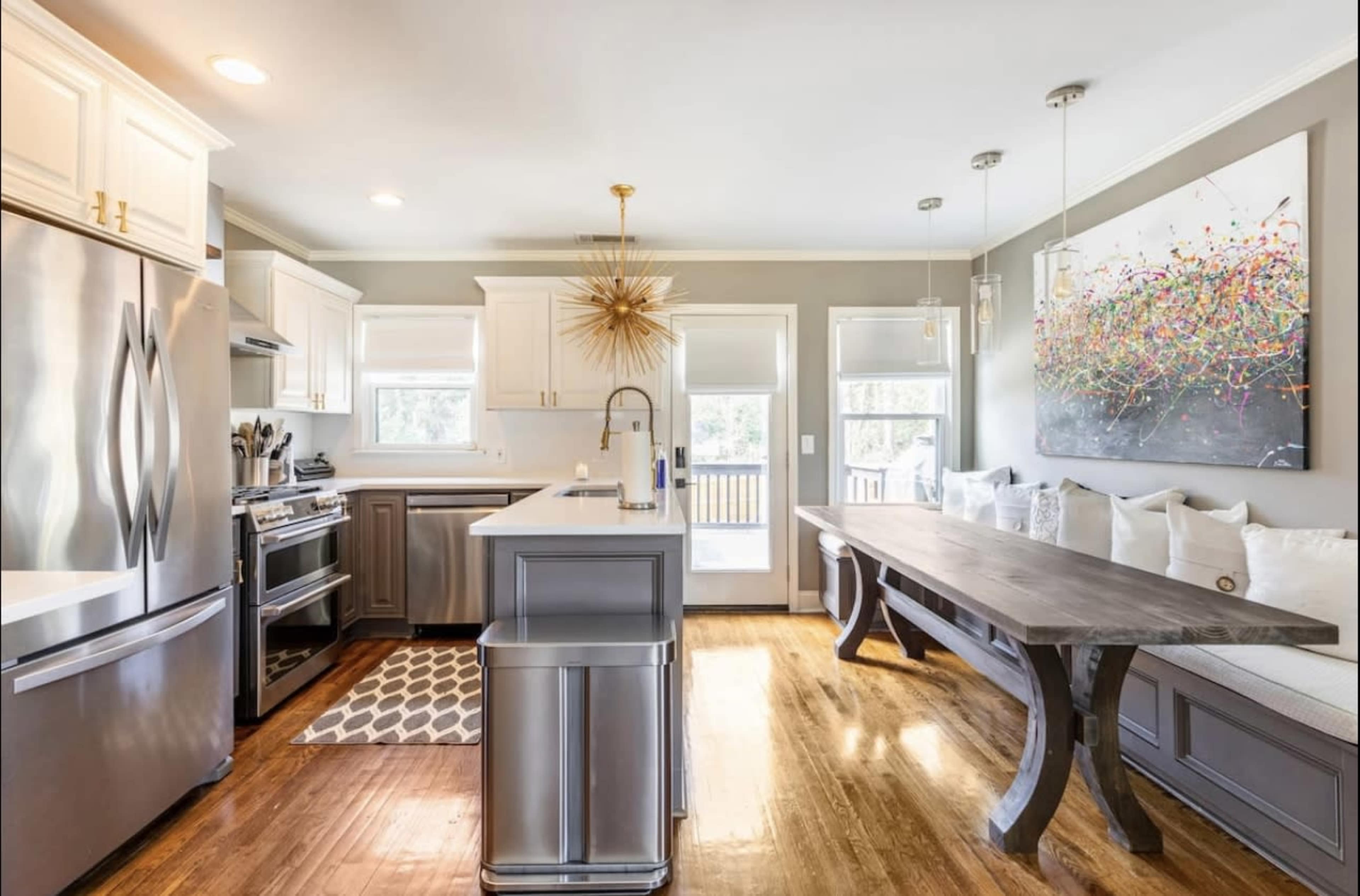 The image depicts a modern kitchen with stainless steel appliances, a large island, and a wooden dining table beside a sunlit window.