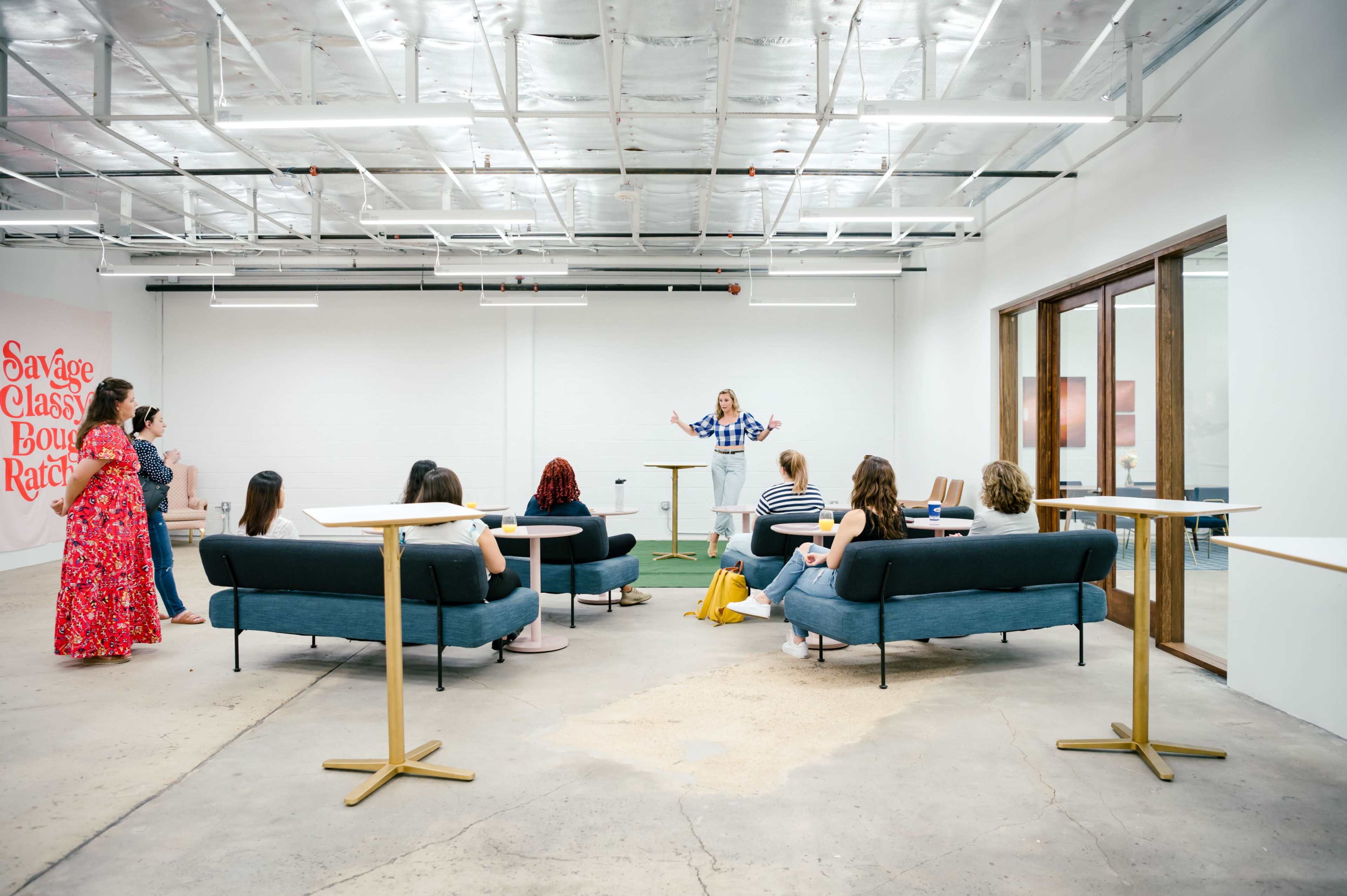 A group of people is seated on couches in a modern, spacious room while a person stands in front of them, speaking.