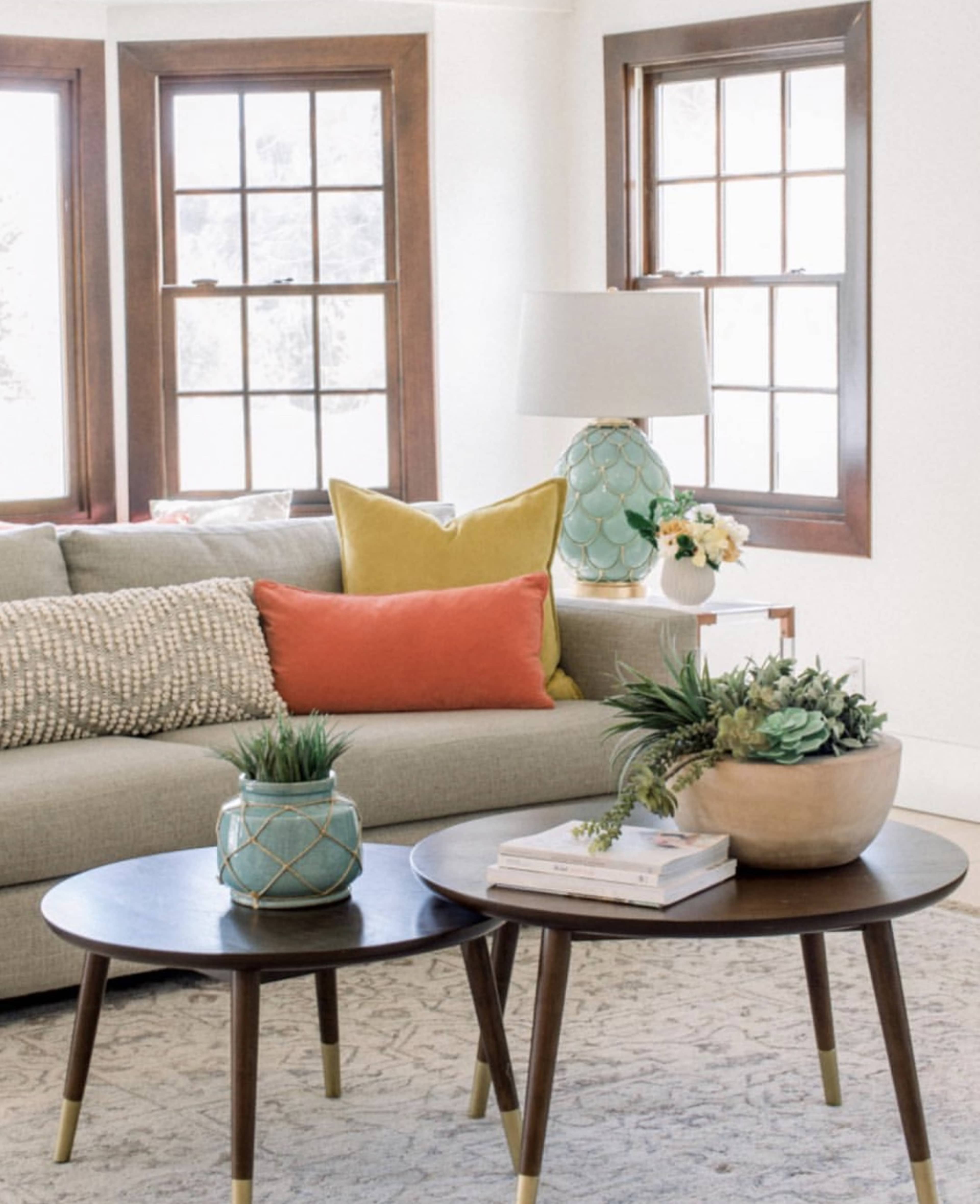 A living room features a gray sofa with colorful cushions, two wooden coffee tables with plants and decor, and large windows allowing natural light.