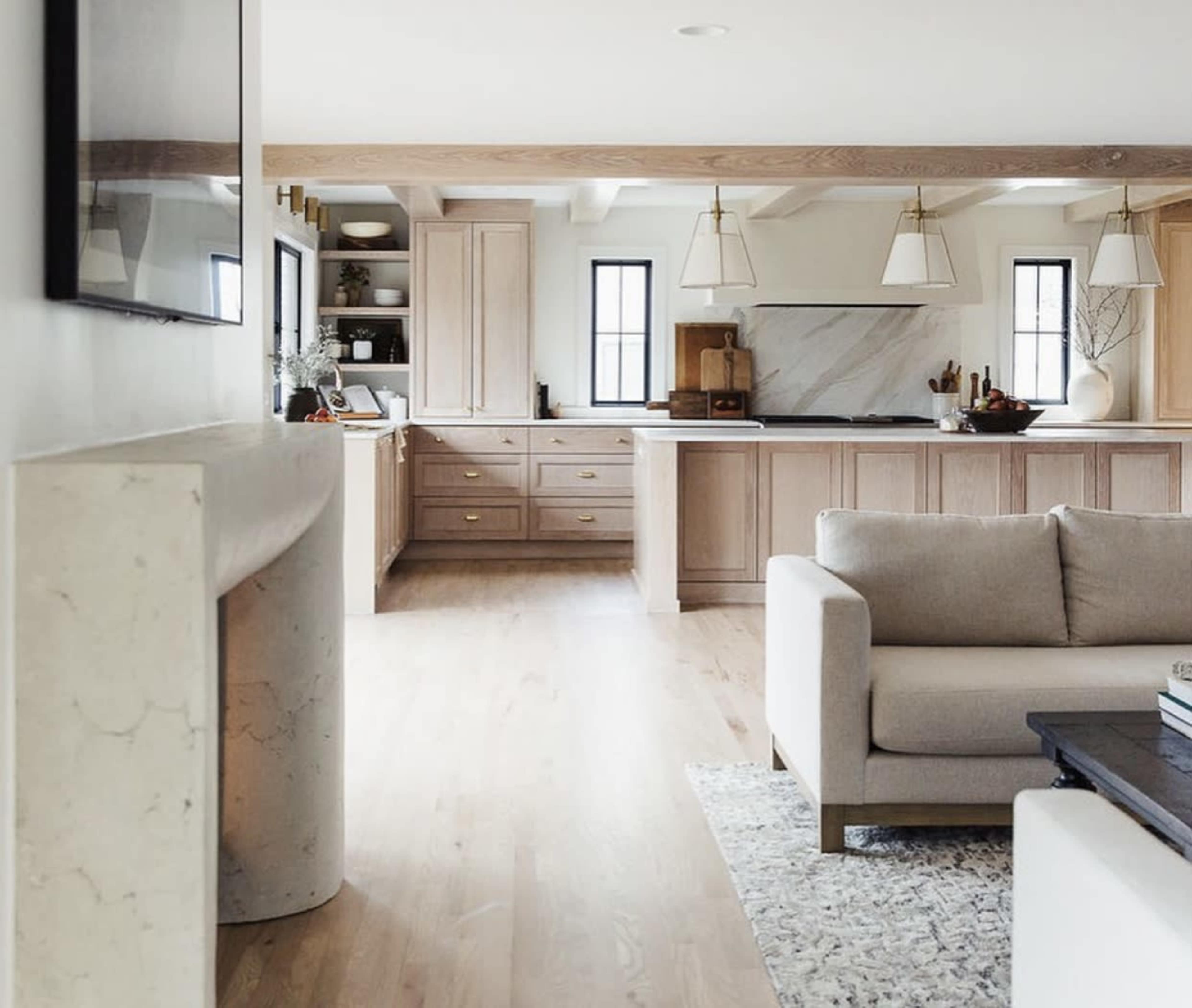 The image shows a modern kitchen and living area featuring light wood cabinets, a spacious countertop, and a cream-colored couch in the foreground.