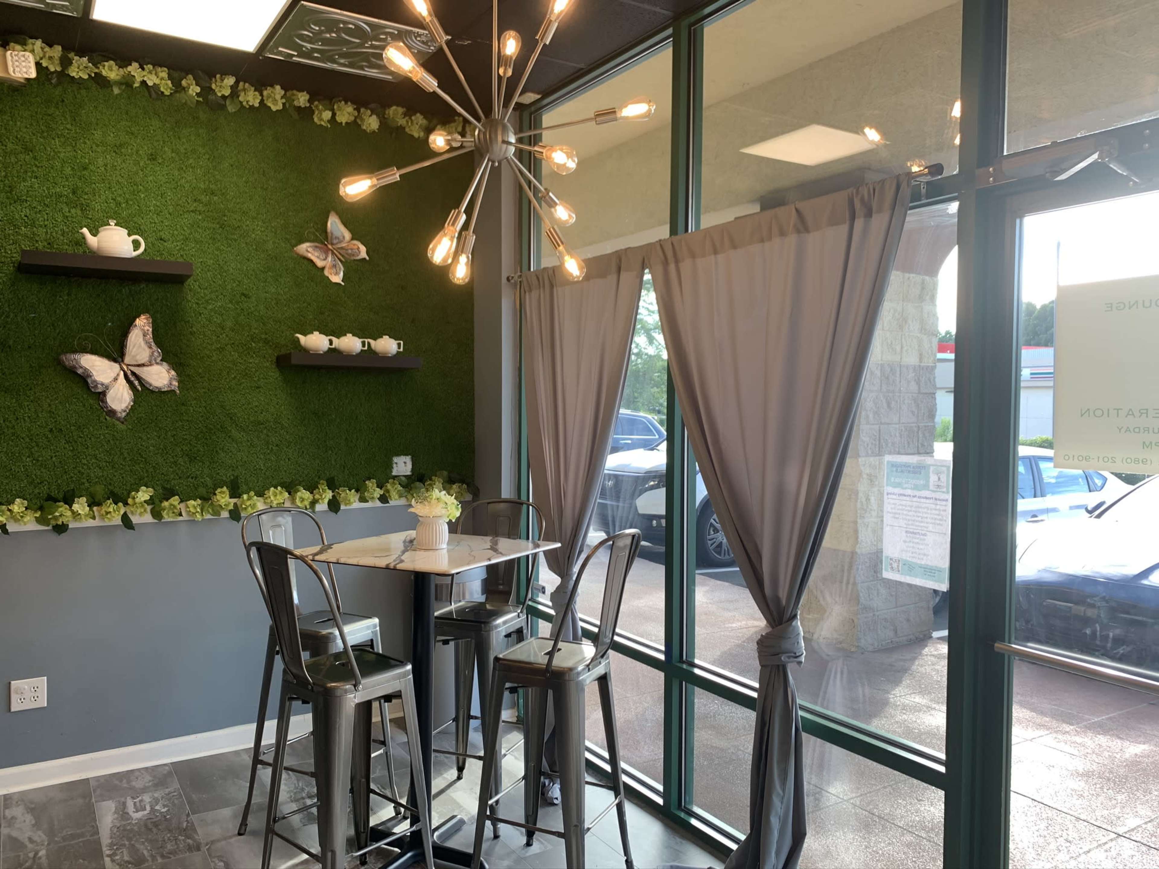 A small cafe interior featuring a green moss wall with decorative butterflies, a table with metal stools, and a large glass entrance with sheer curtains.