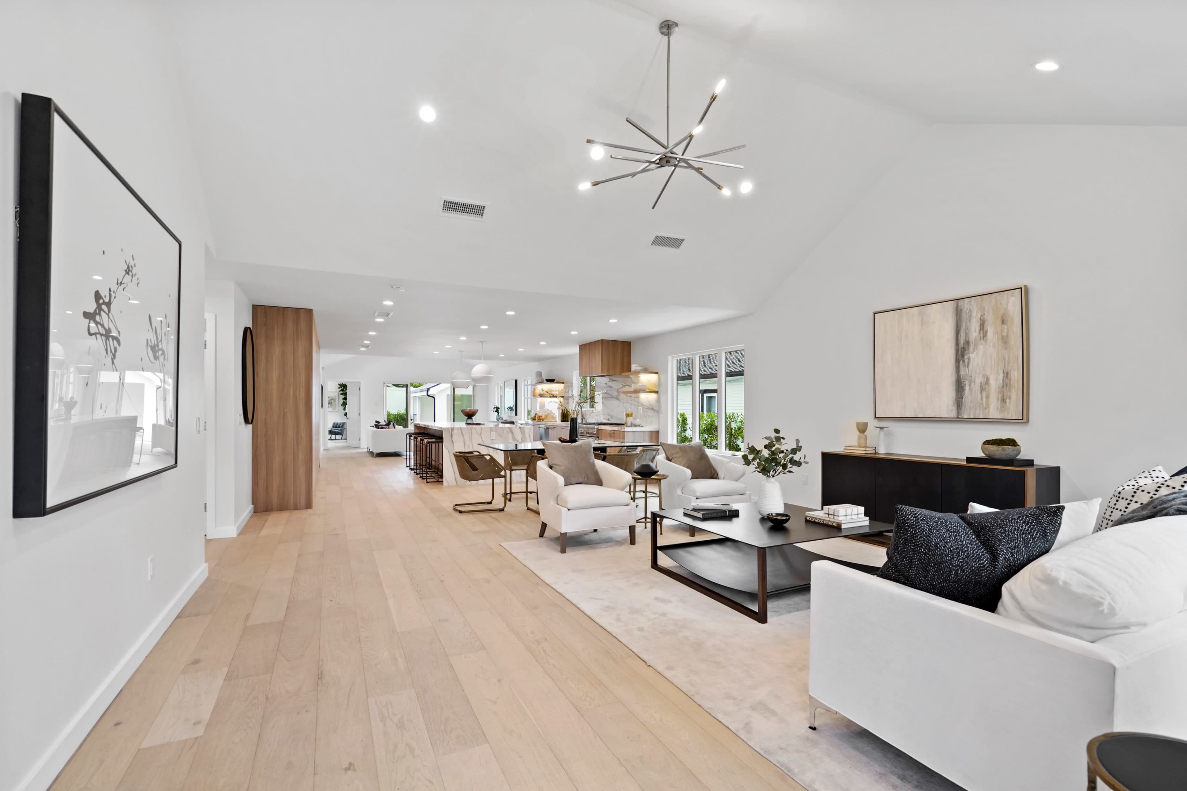 A spacious, modern living room with light wood floors, high ceilings, and contemporary furniture, leading into a kitchen area.