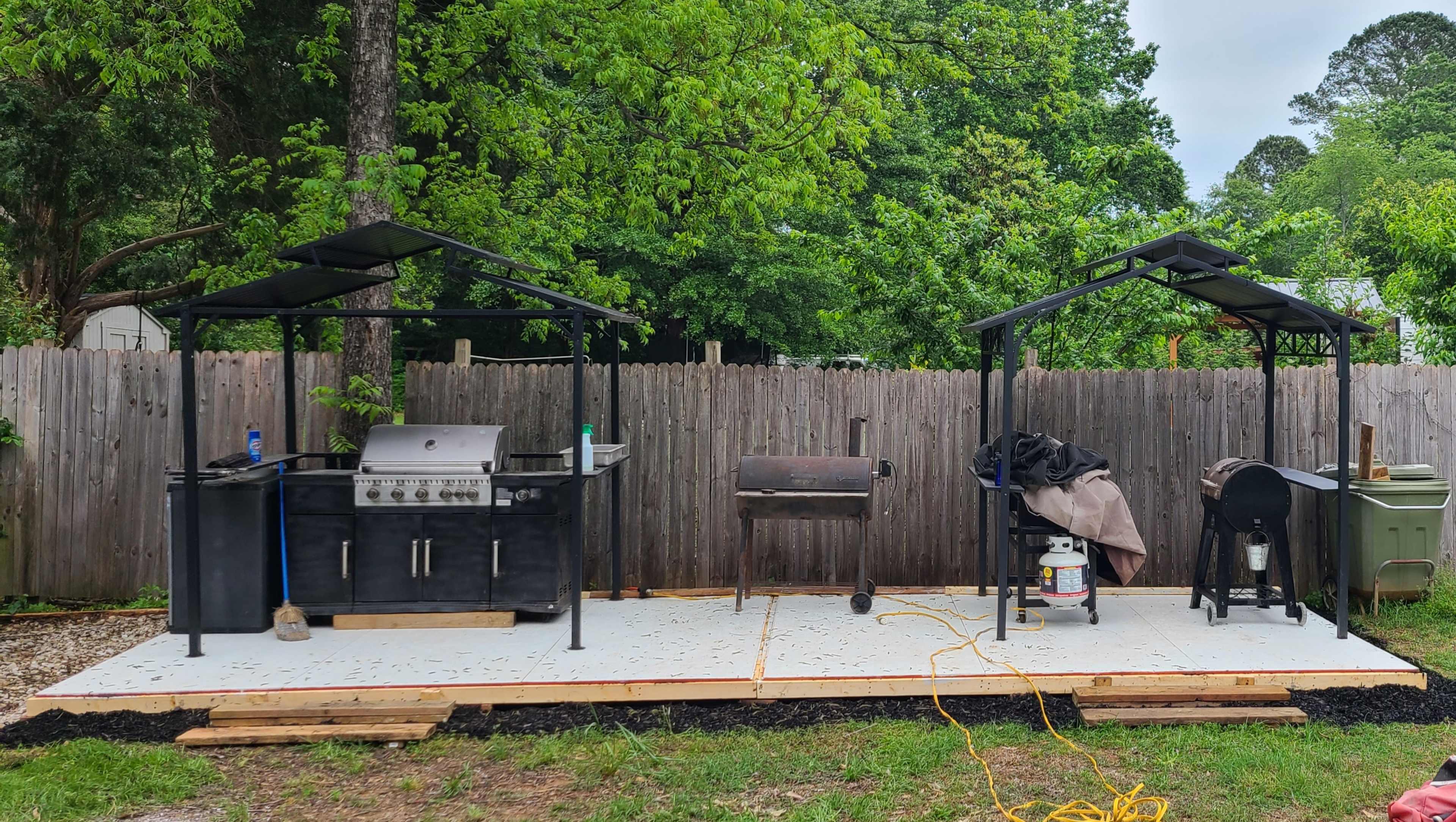 An outdoor grilling area with a black gas grill and a charcoal smoker under separate covers, situated on a concrete slab surrounded by landscaping.